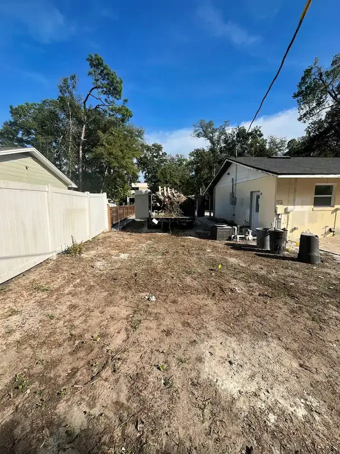 Backyard with dirt ground, a white fence, and two houses. Construction debris visible. Blue sky.