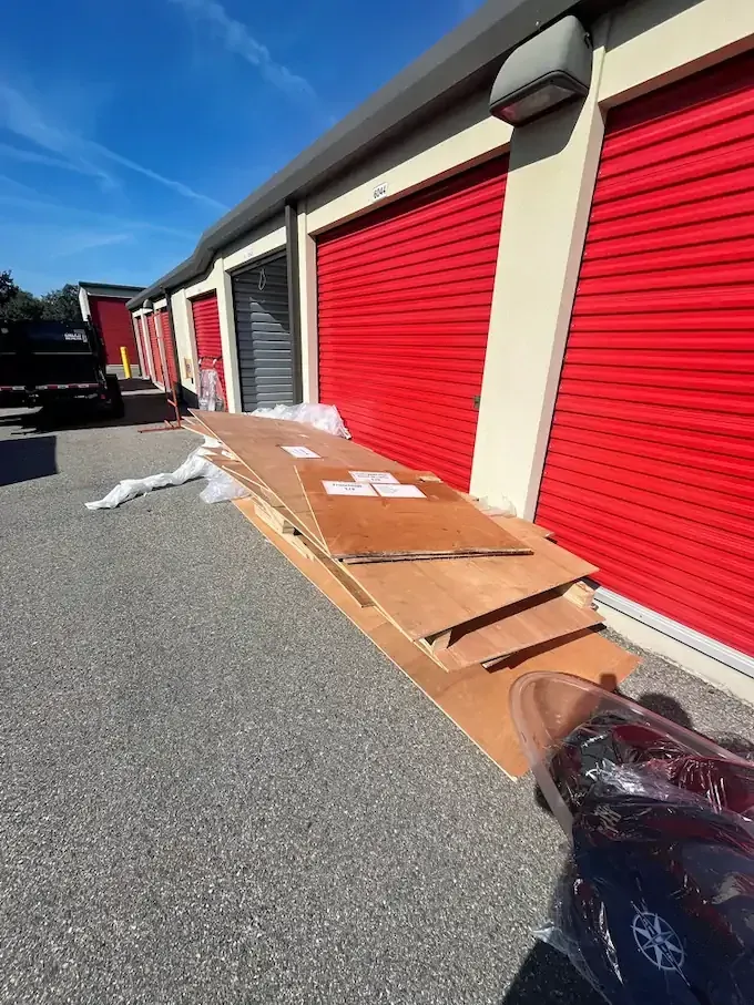 Plywood boards on gravel outside storage units with red doors under a blue sky.