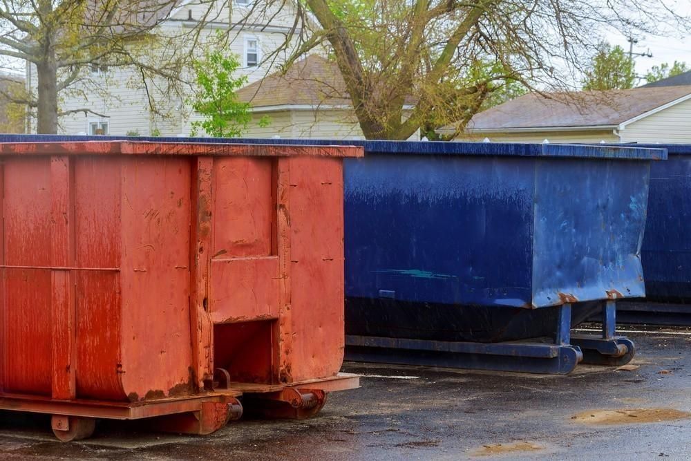 Red and blue dumpsters sit on a paved lot, likely for waste disposal.