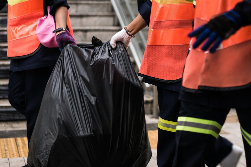 Three people in safety vests carrying a black garbage bag near stairs.