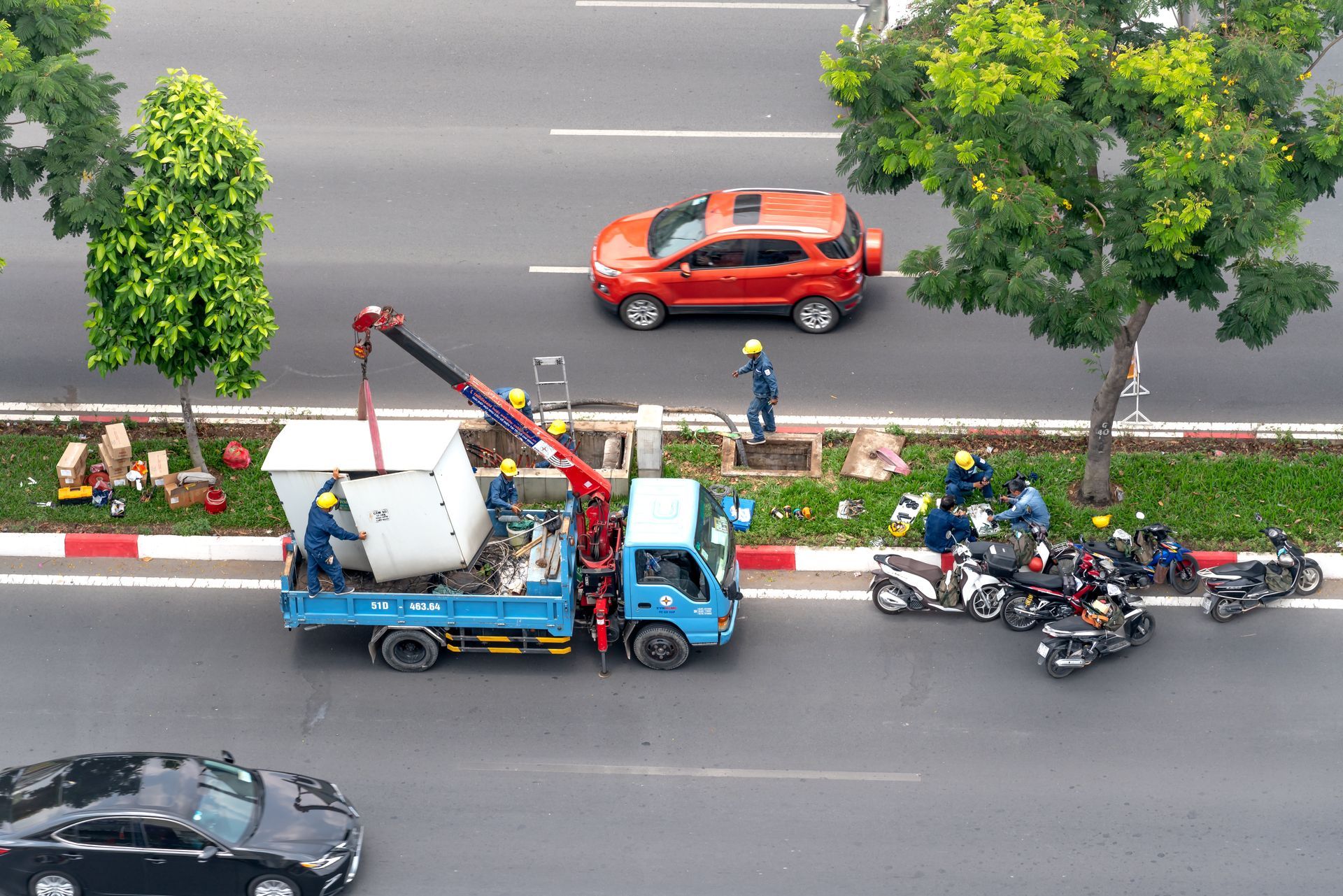 Workers using a crane to lift a large white object from a truck on a city street.