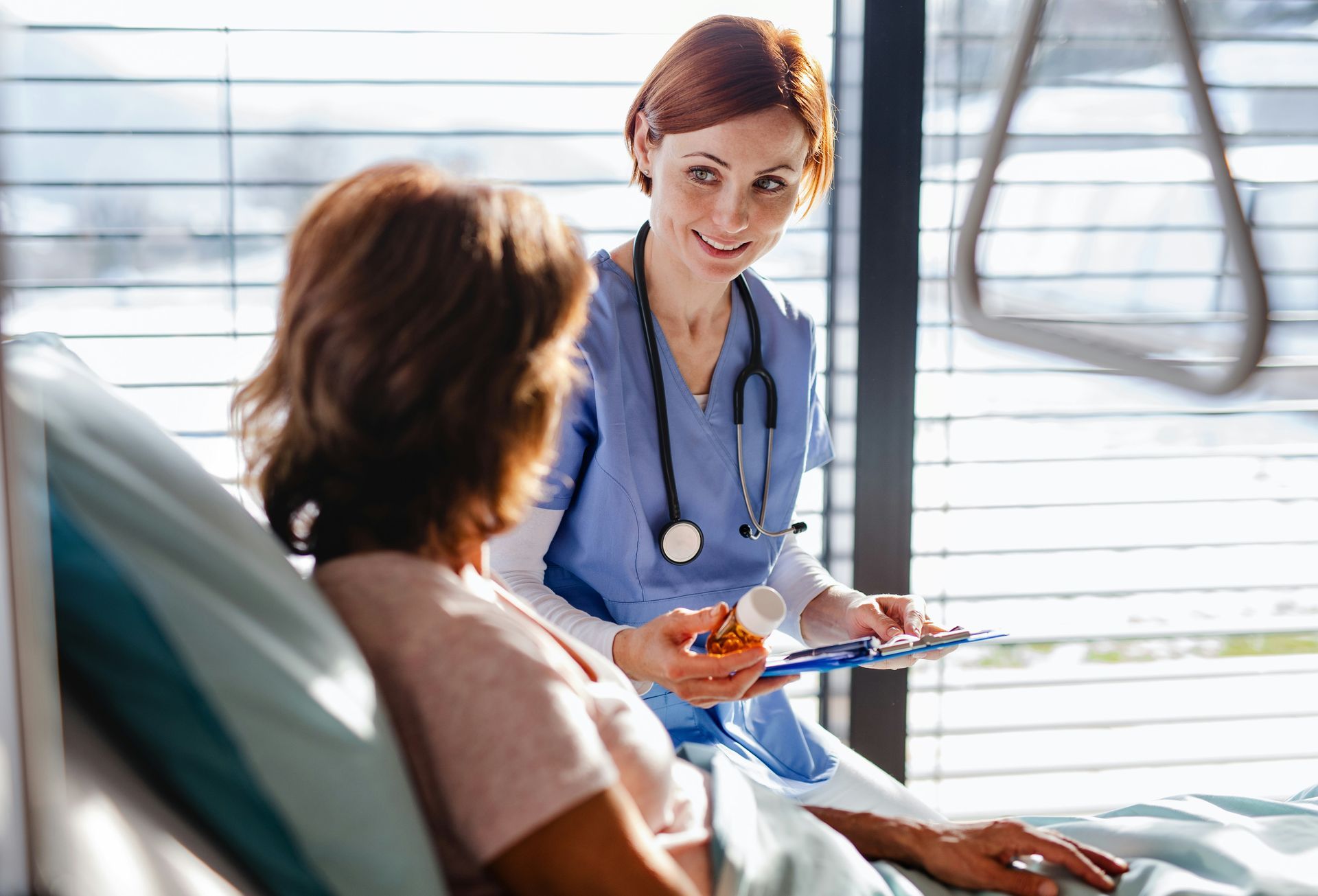 Nurse in blue scrubs speaking with a patient in a bright hospital room, holding a clipboard and pen
