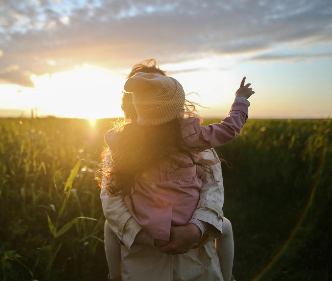 Child on an adult’s shoulders in a field, reaching toward a glowing sunset.