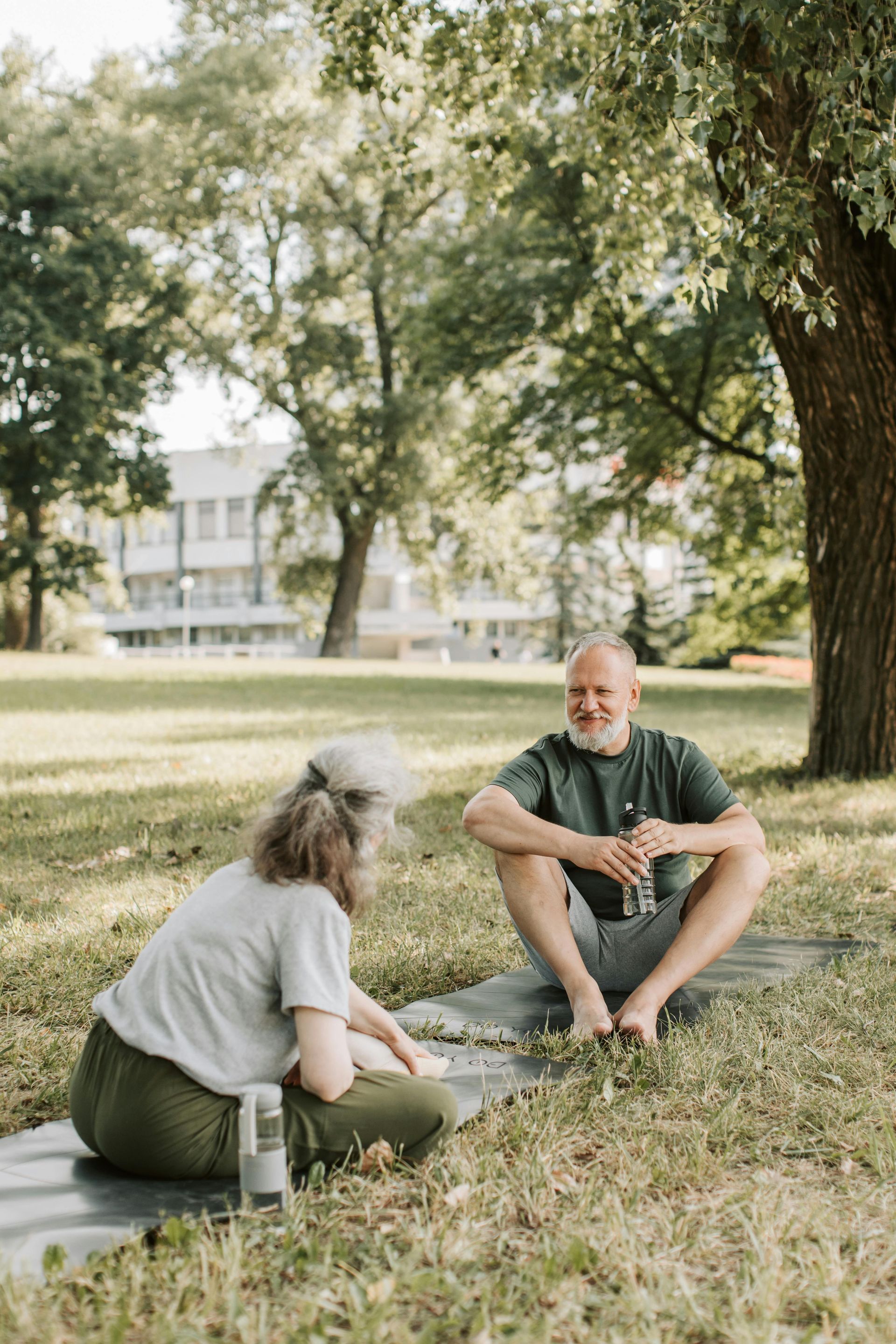 Two people sitting on a blanket in a park under trees, talking in a sunny grassy area.
