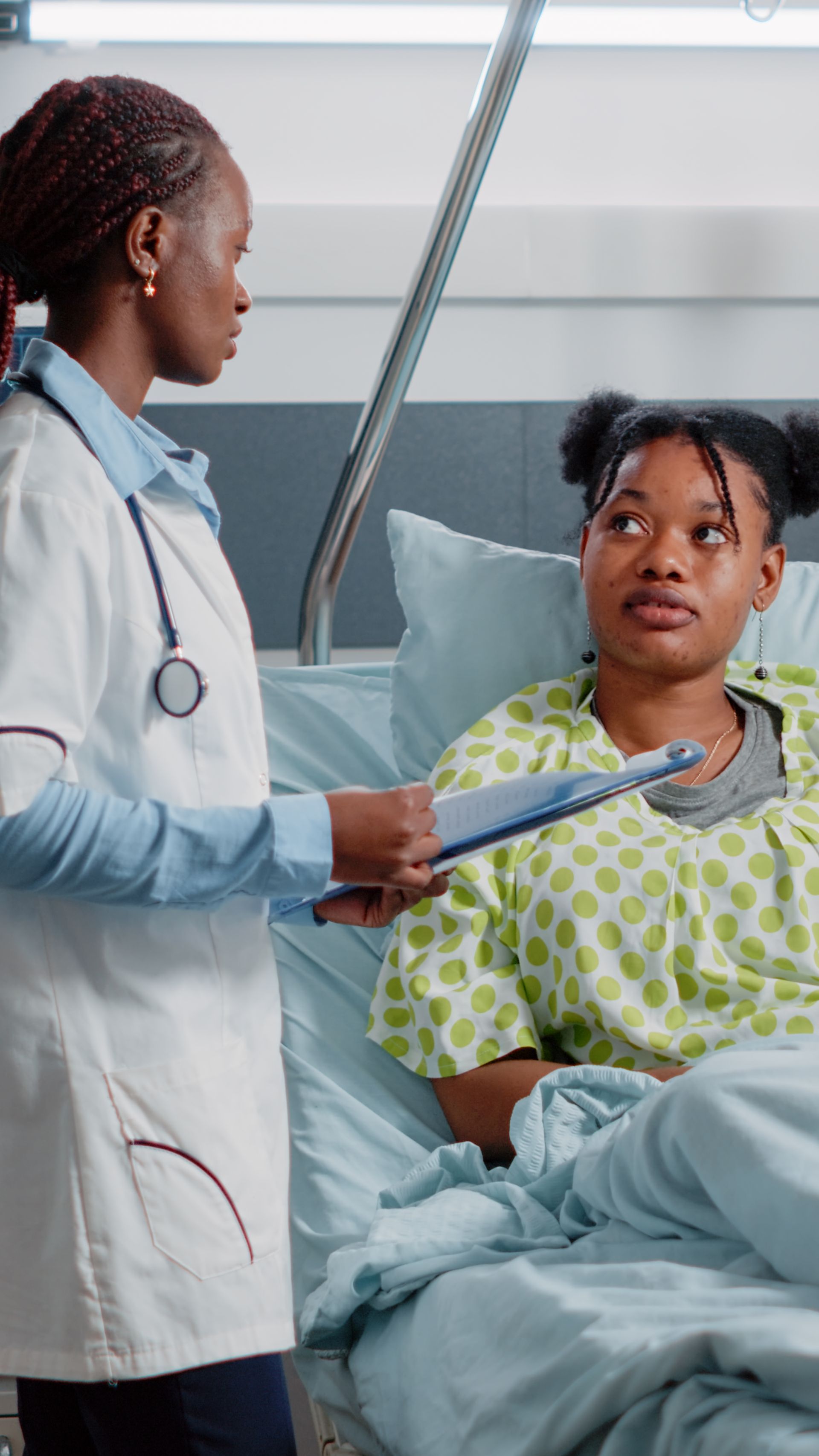 Doctor speaking with hospital patient in bed, holding clipboard in a bright clinic room