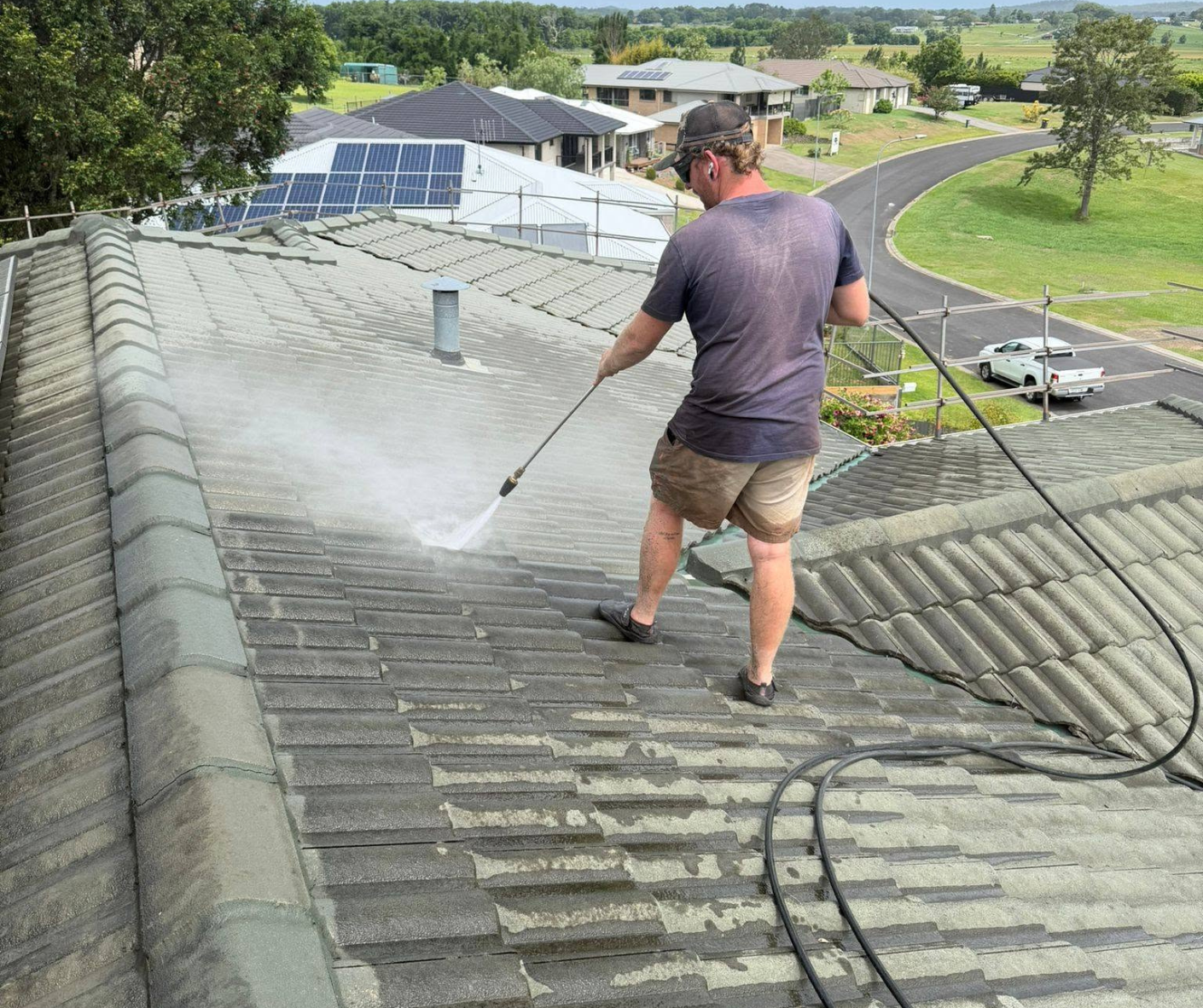 Worker pressure-washing a gray tiled roof on a sunny suburban home Worker pressure-washing a gray tiled roof on a sunny suburban home