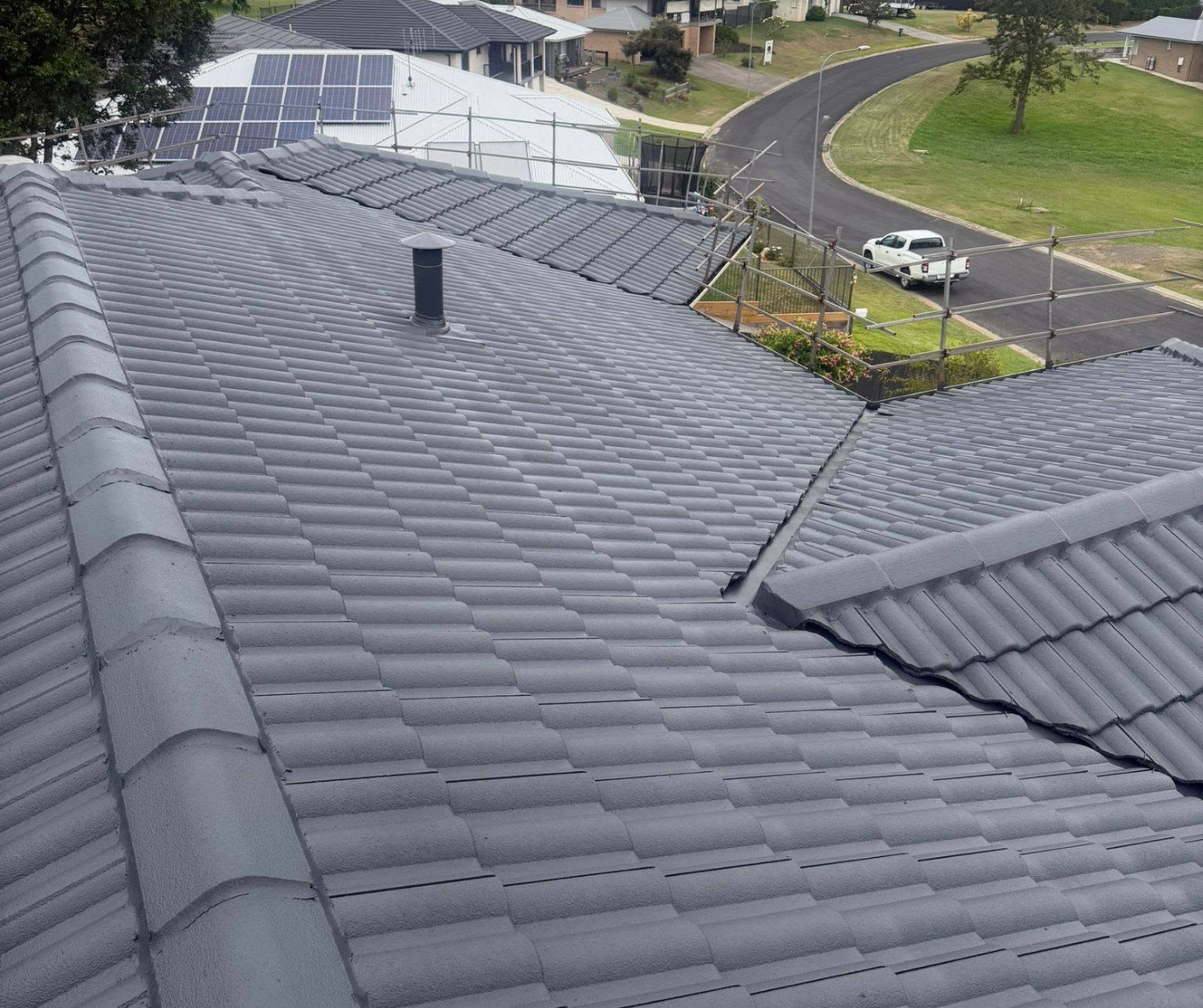 Roof with dark gray tiles, solar panels, and a vent, viewed over a suburban street and houses. Roof with dark gray tiles, solar panels, and a vent, viewed over a suburban street and houses.