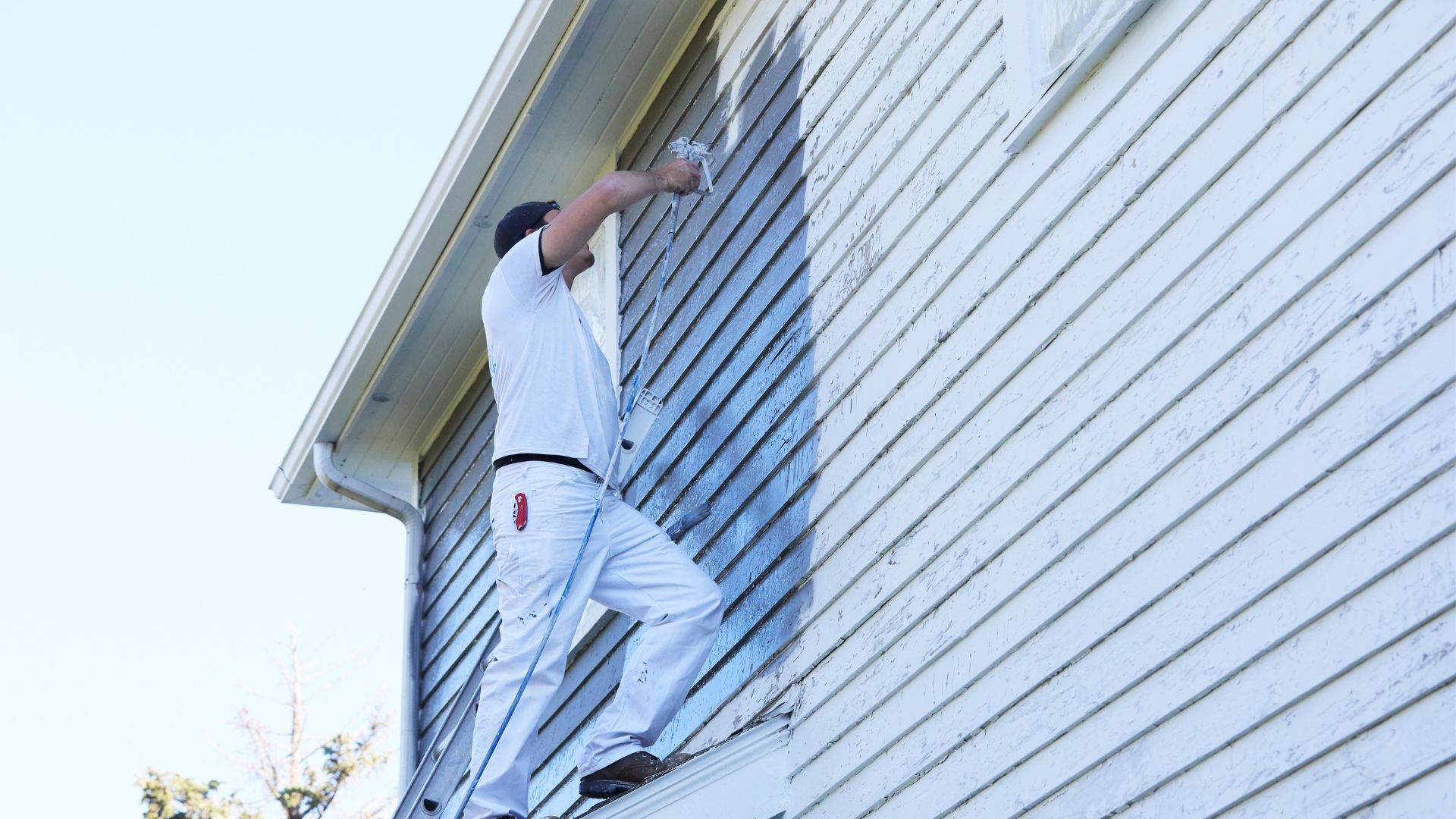 Person on a ladder painting the upper exterior siding of a house.