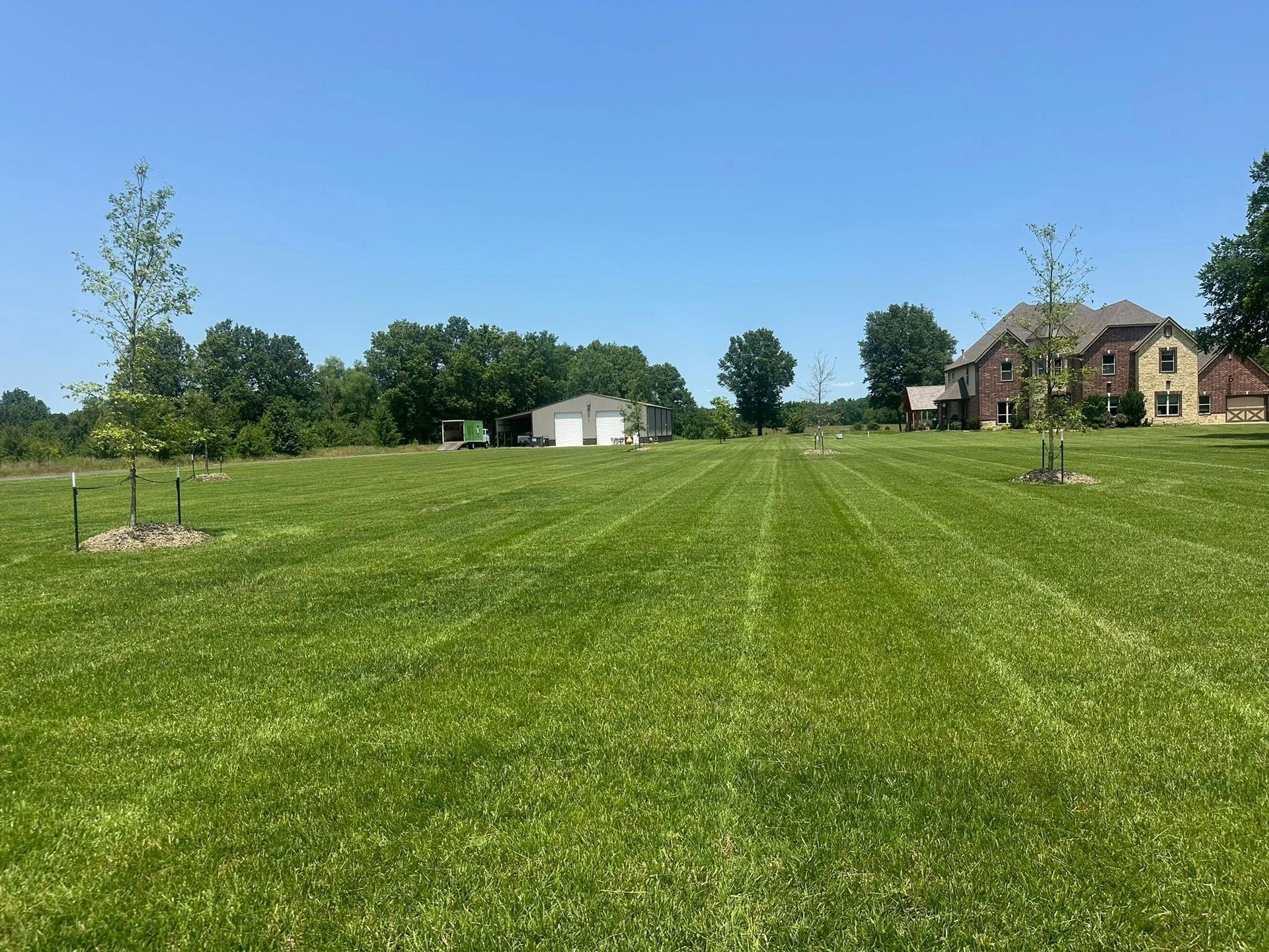 Lush green lawn with mowed paths, small trees, and a house with a garage under a clear blue sky.