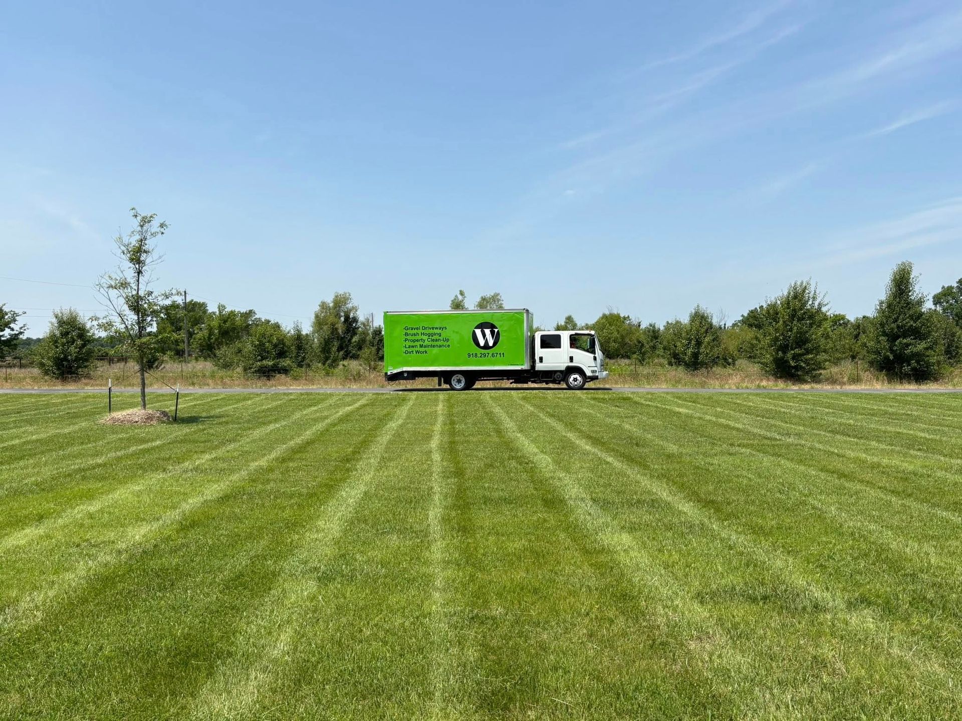 Green truck with logo on grass field, sunny day, trees in background.