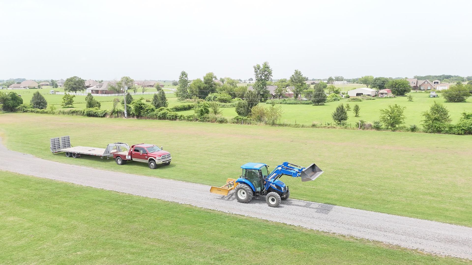 A blue tractor grading a gravel driveway, with a red truck and trailer parked nearby in a grassy field.