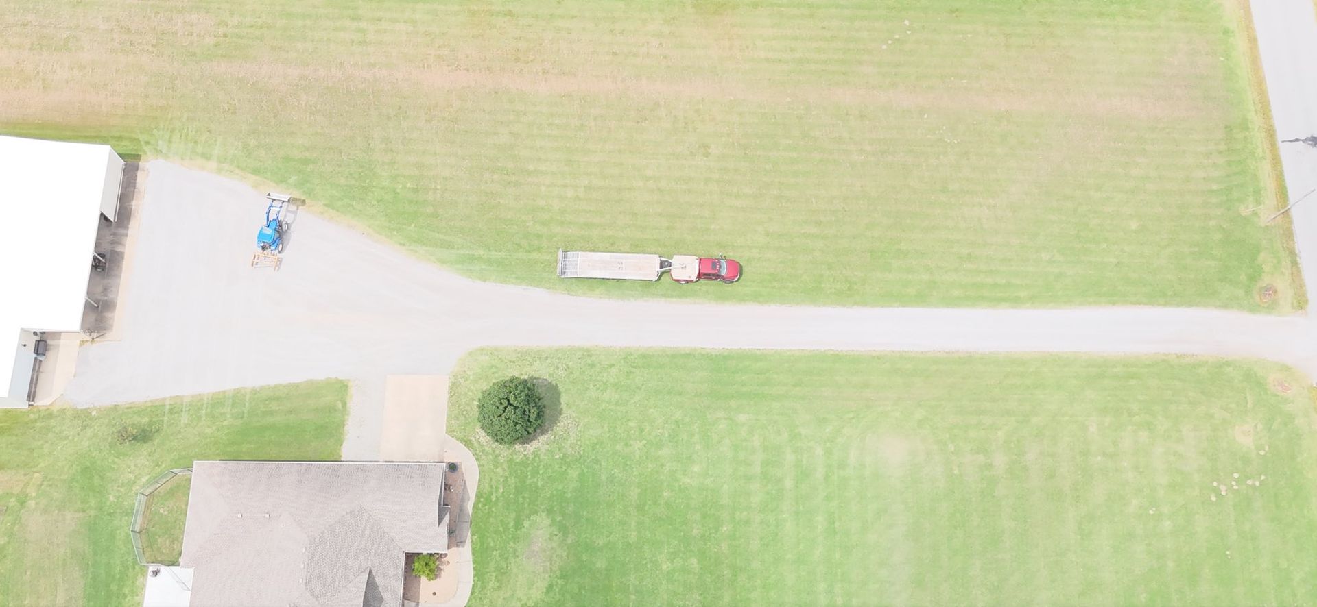 Aerial view of a farm scene: a tractor pulling a trailer on a gravel road, surrounded by fields and a building.