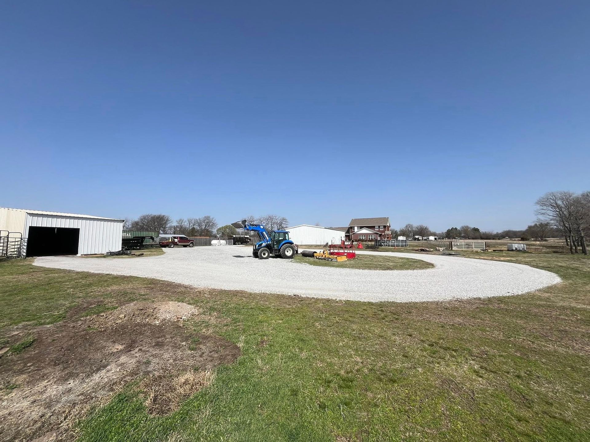 Gravel driveway curves through grassy field, leading to buildings under a blue sky. A tractor sits mid-driveway.