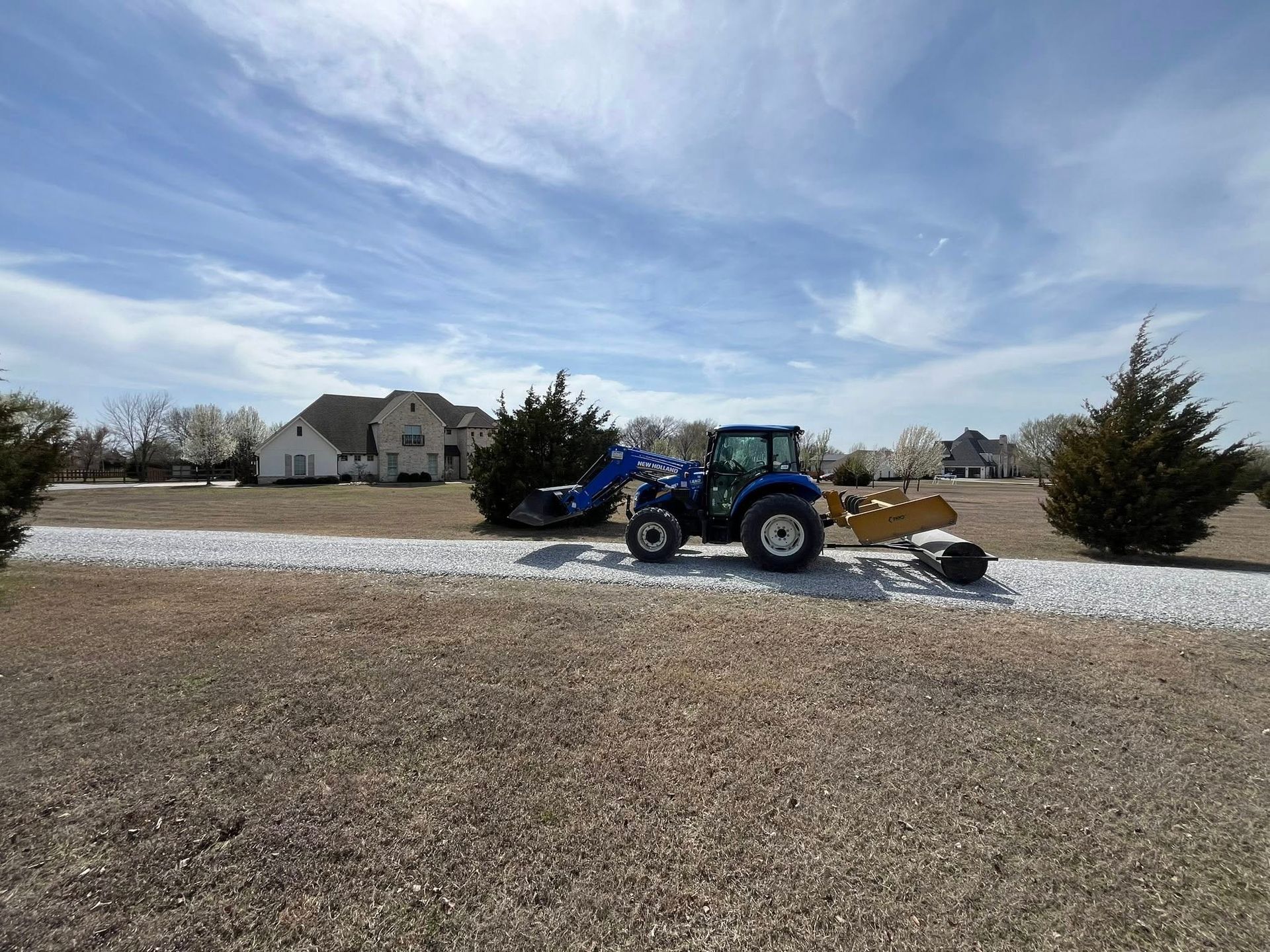Blue tractor grading a gravel driveway in front of houses under a partly cloudy sky.