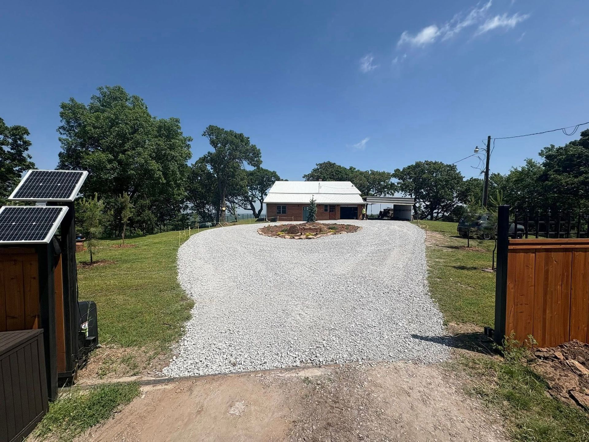 Gravel driveway leading to a light-colored house under a blue sky, flanked by trees and solar panels.