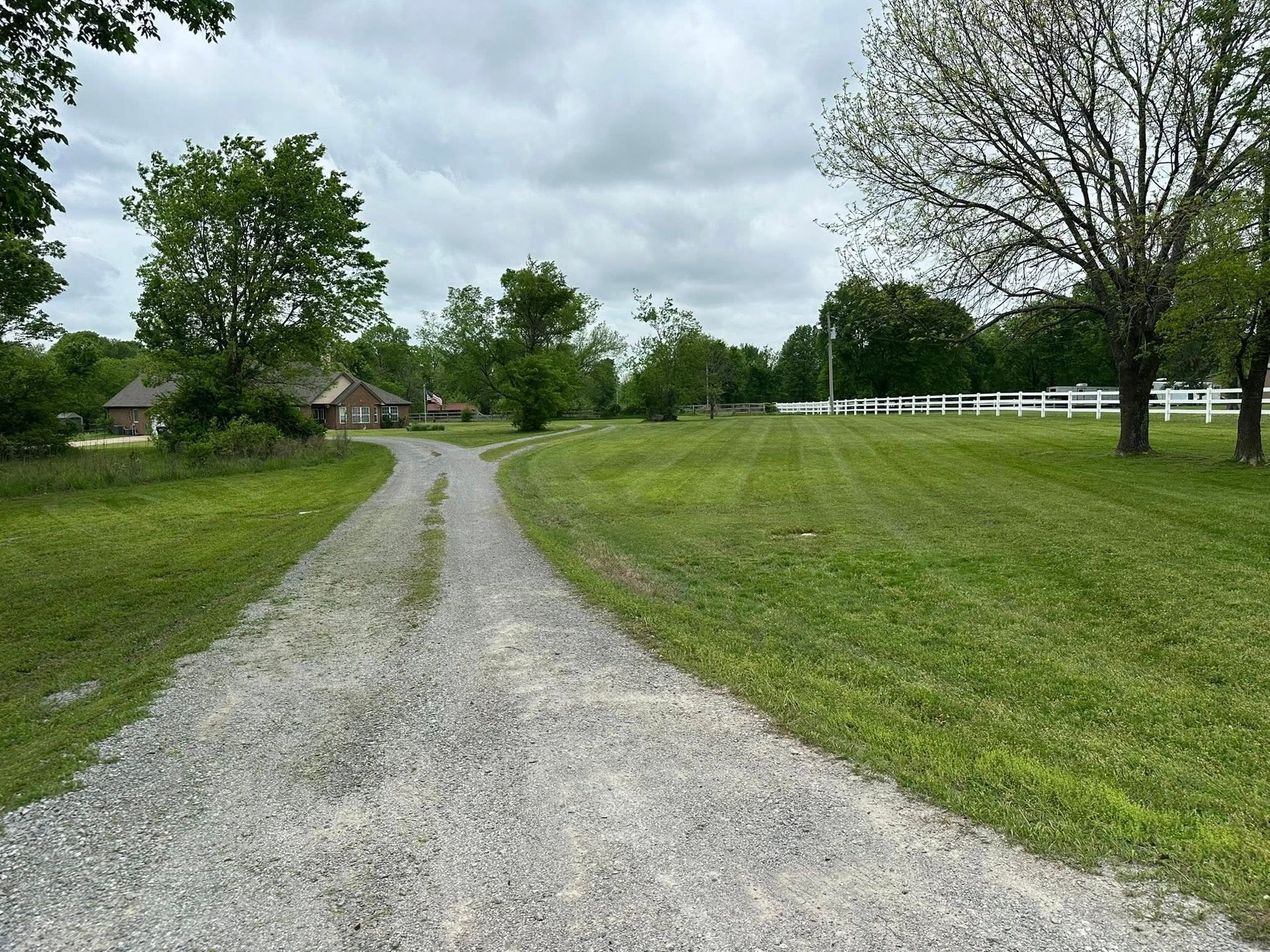 A gravel driveway leads to a brick house on a green lawn, overcast sky.