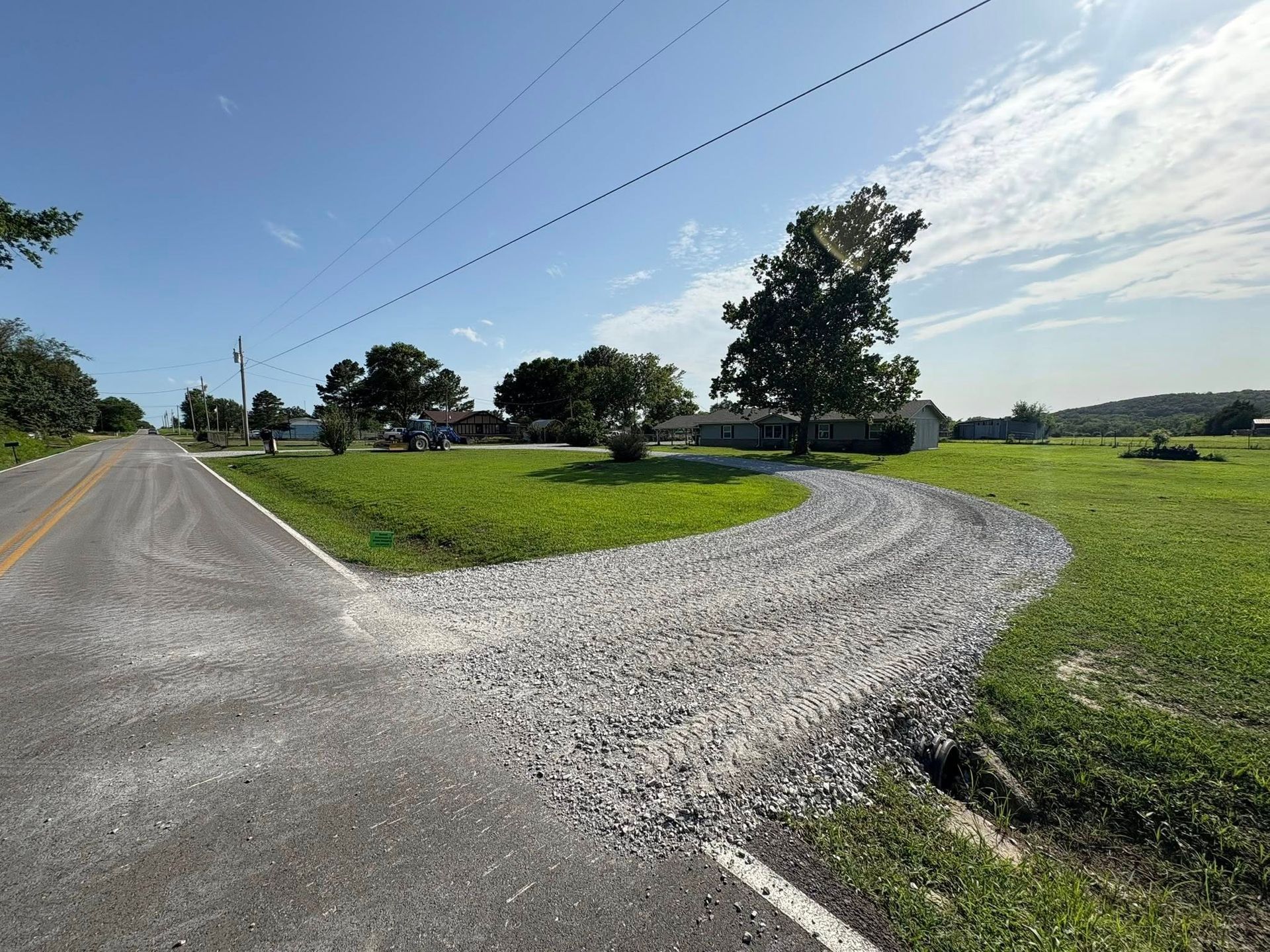 Gravel driveway curves from paved road into grassy area with trees and blue sky.