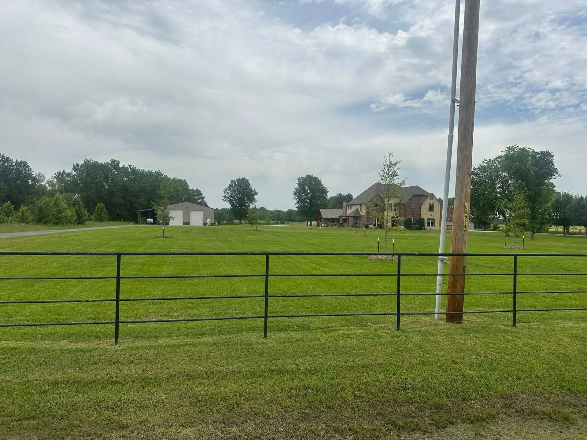 Green field with a black fence, a house, and a utility pole under a cloudy sky.