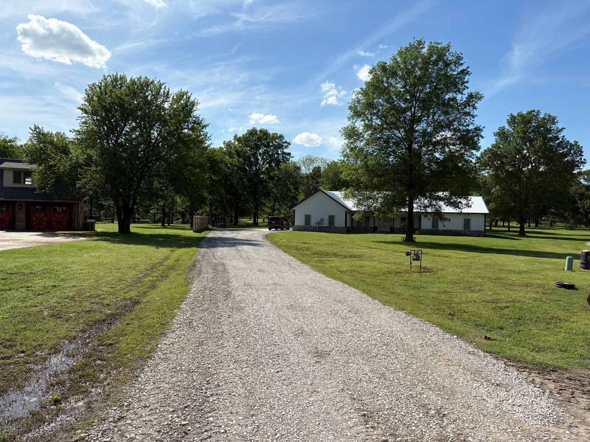 A gravel driveway leads to a white house under a blue sky, flanked by green grass and trees.