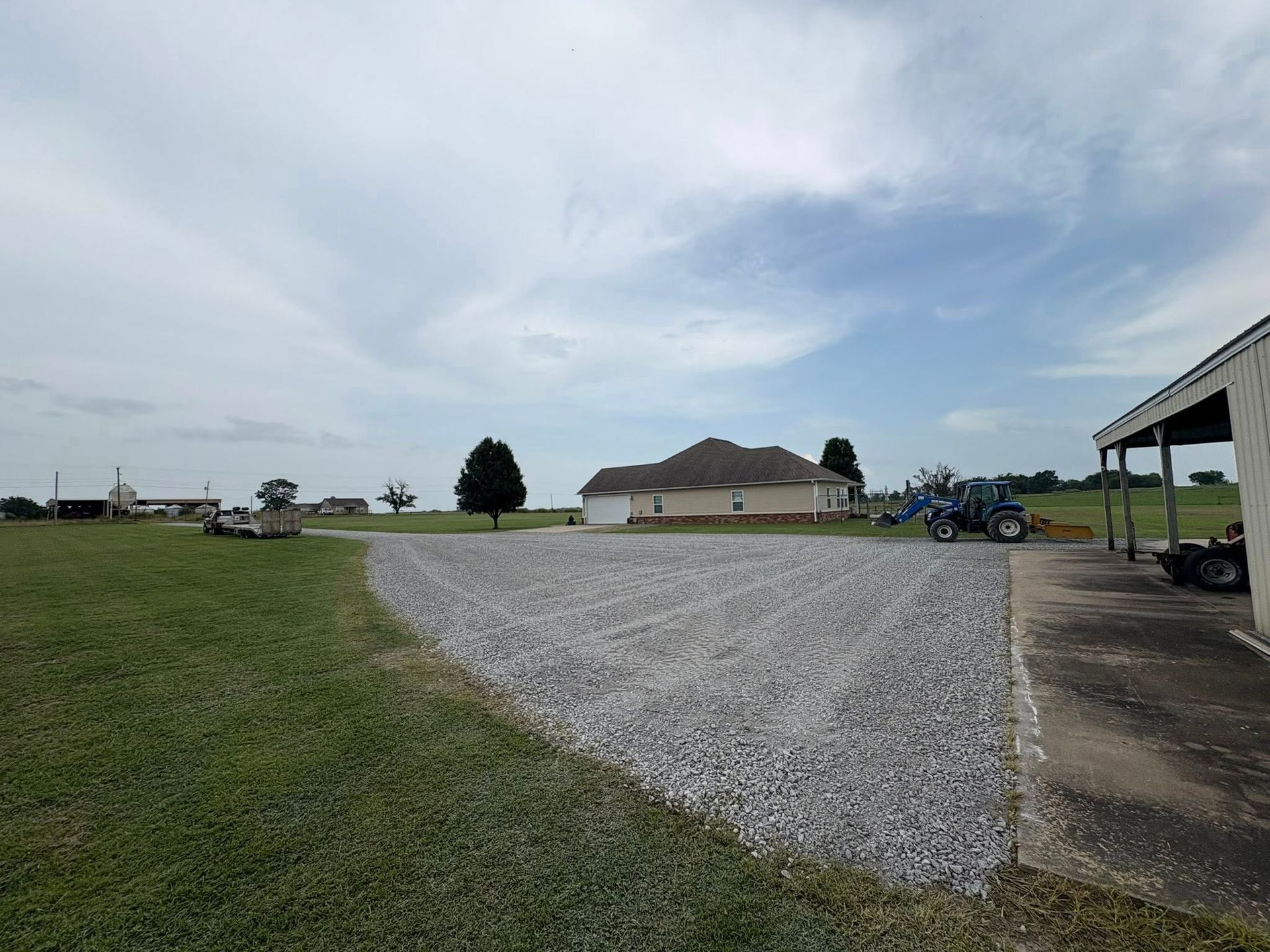 A gravel driveway extends towards a house under a cloudy sky. A tractor sits nearby.