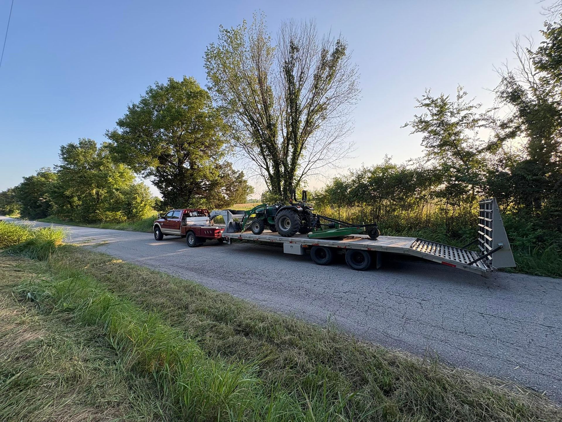 Red truck pulling a trailer with a green tractor on a gravel road, trees in background.