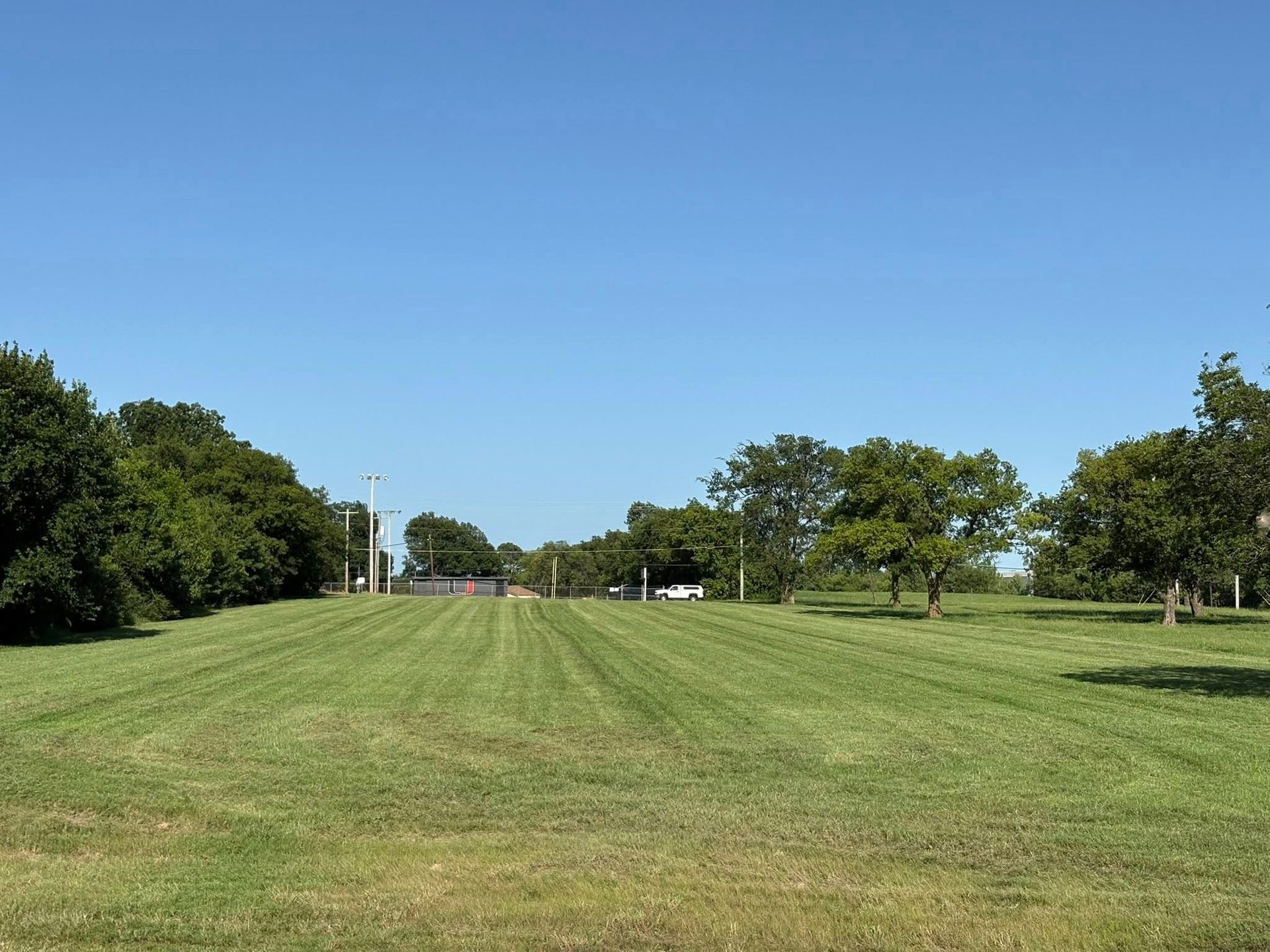 Mowed green field under a clear blue sky, with trees lining the sides and a distant structure.