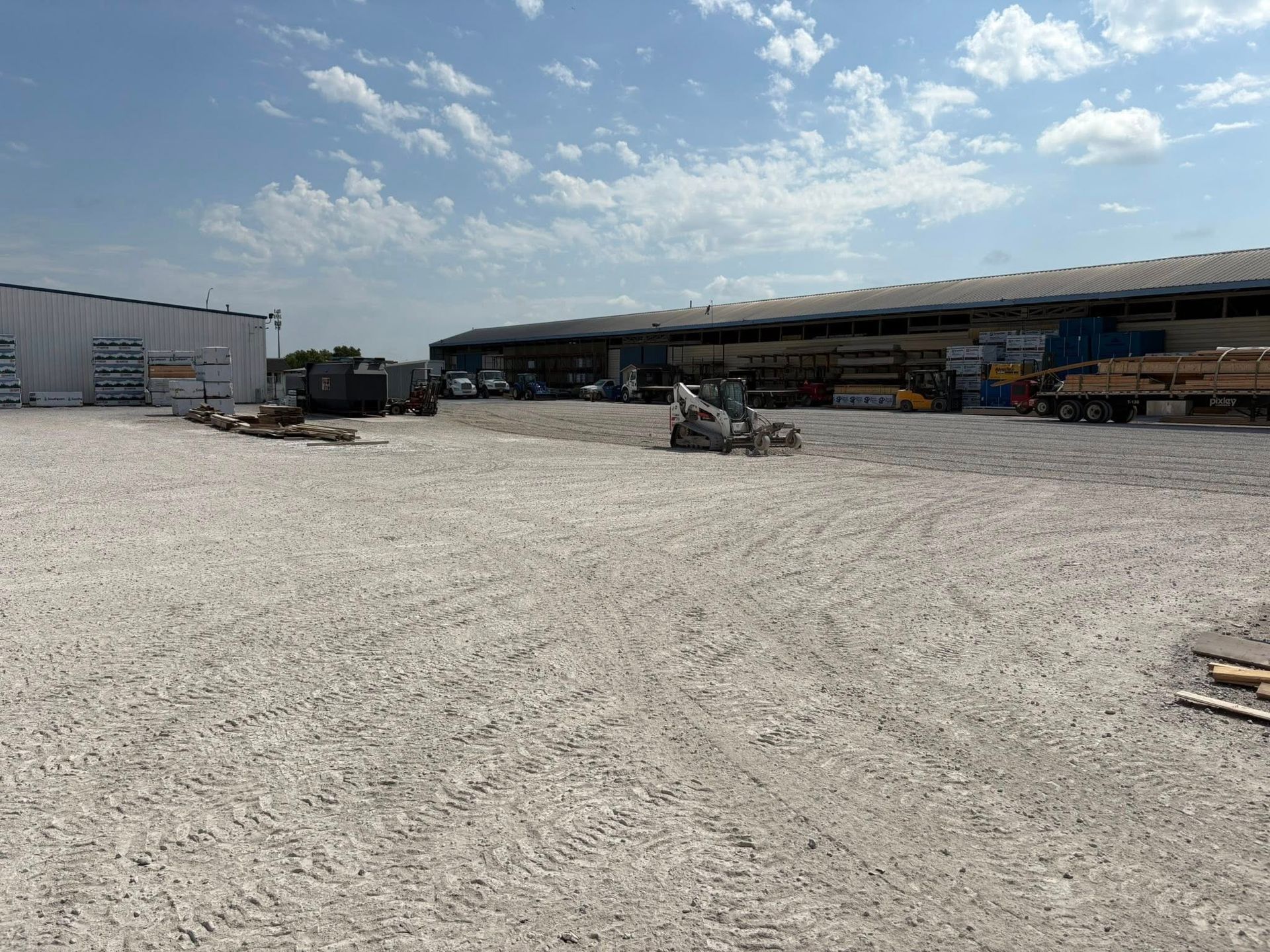 Gravel yard with construction materials and buildings under a partly cloudy sky.