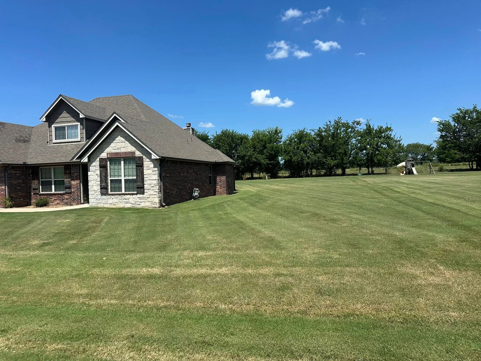 House with brown brick and gray roof; freshly mowed green lawn; blue sky with clouds.
