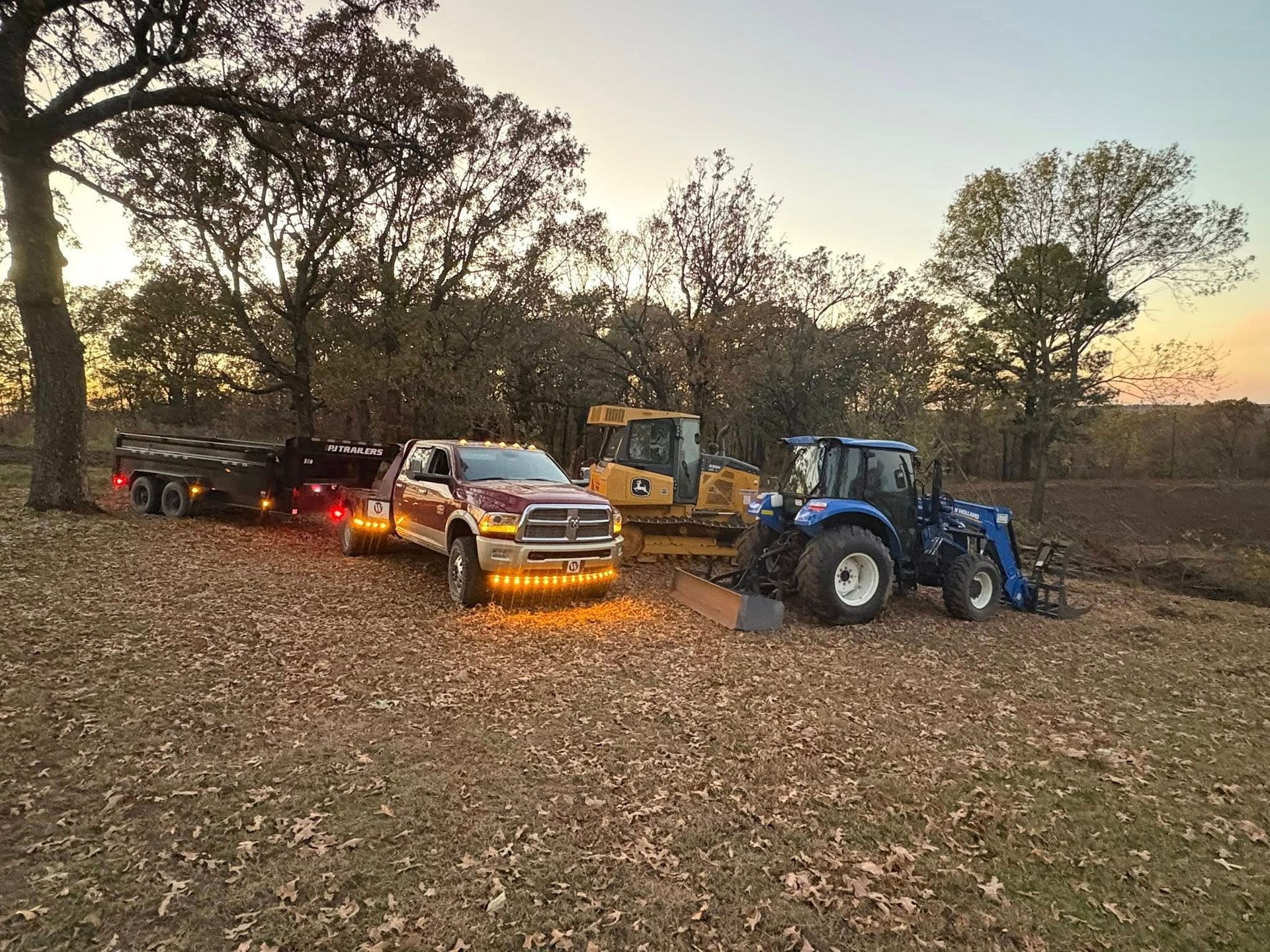 A pickup truck towing a trailer with a bulldozer and tractor in a field at dusk.