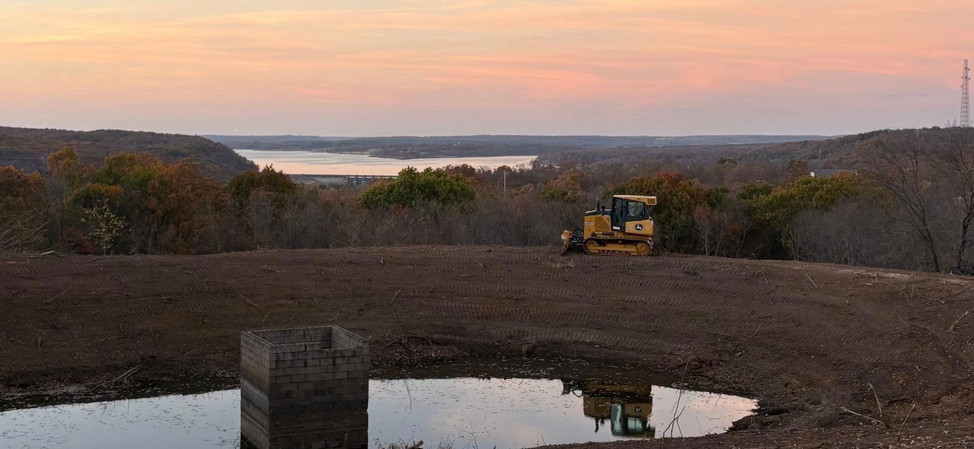 A bulldozer on a barren hilltop overlooking a lake at dusk. A tower is visible in the distance.