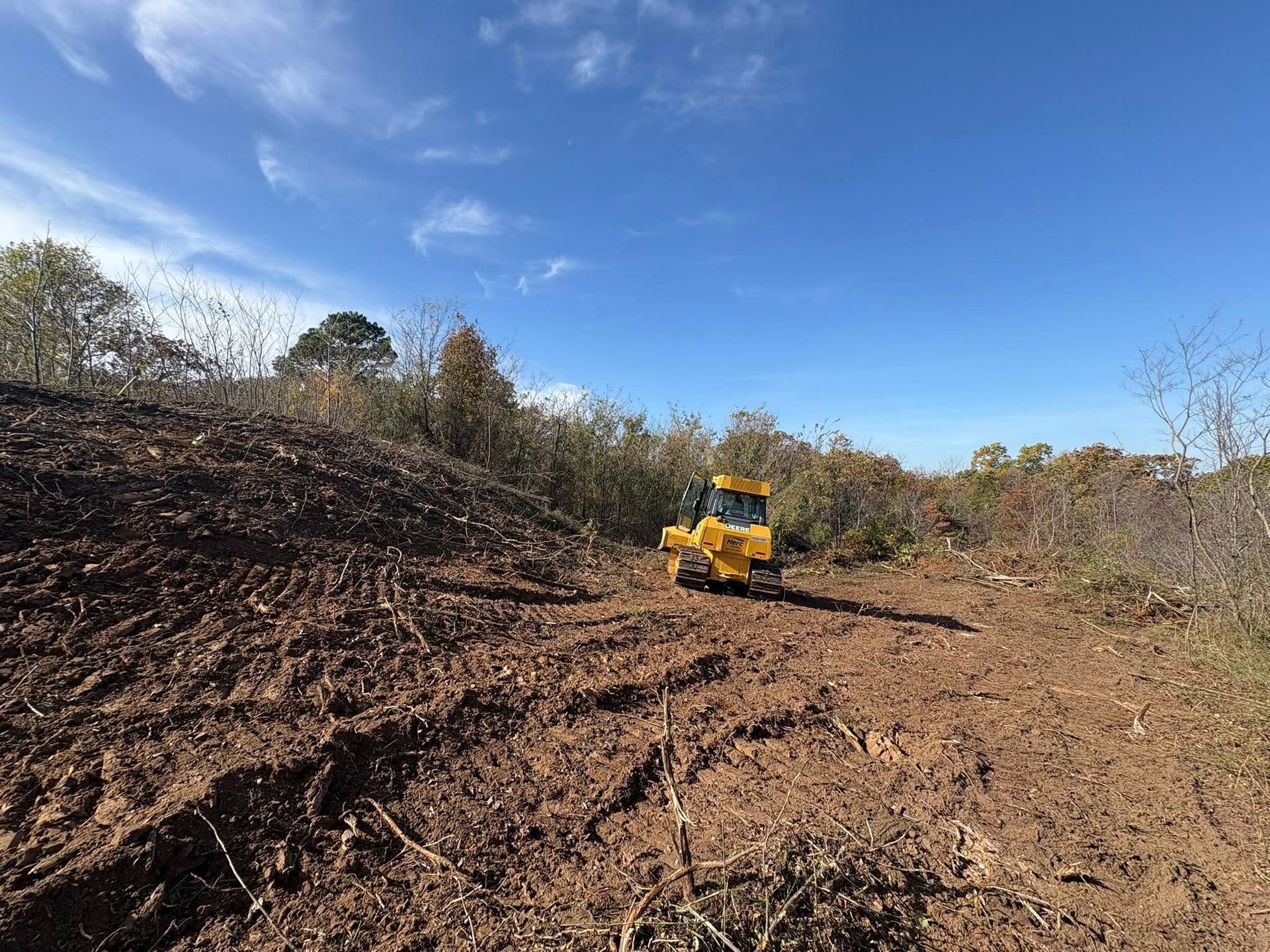 A yellow skid steer tractor on a dirt path in a wooded area with a clear blue sky.
