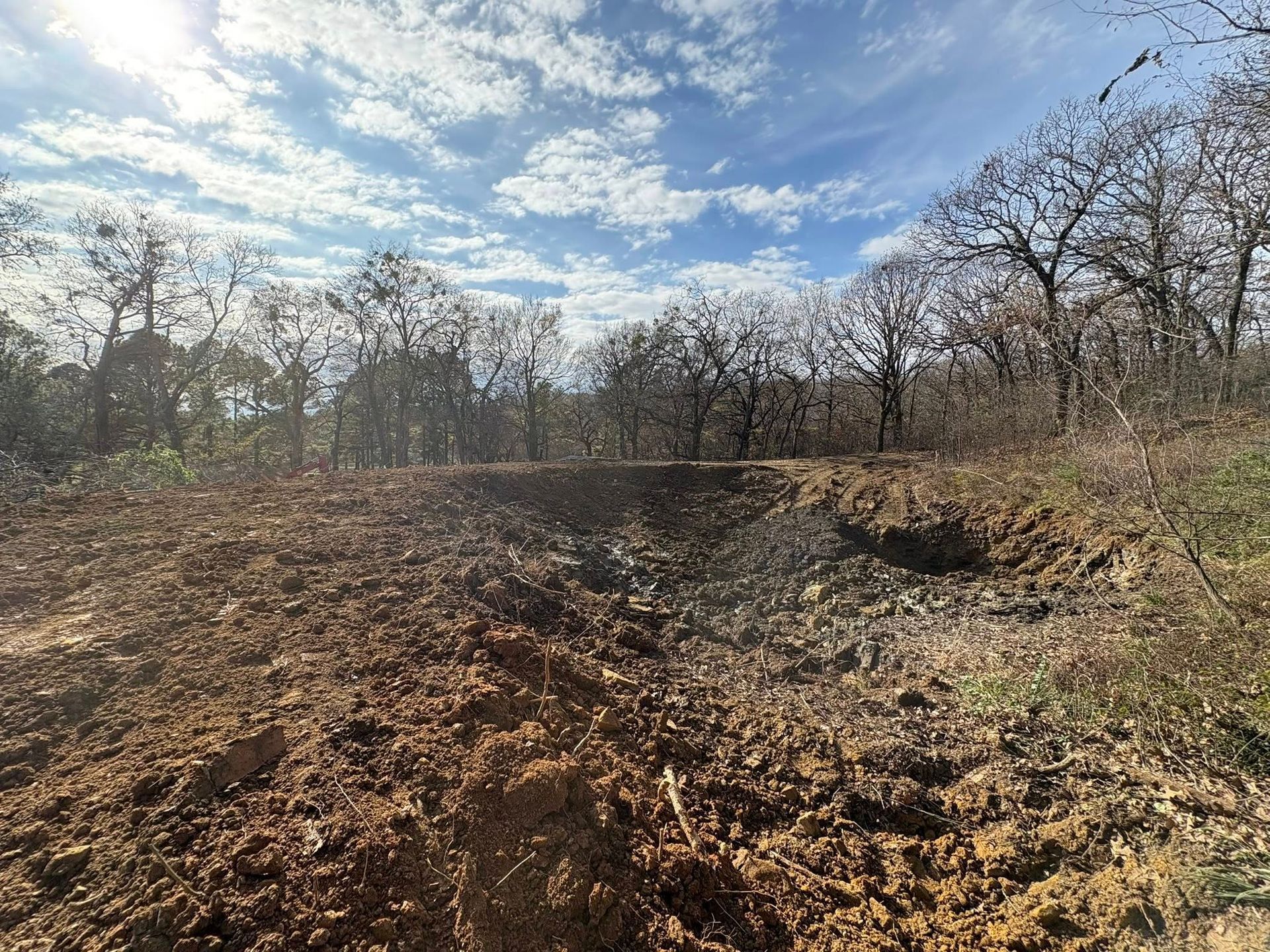Cleared brown earth with a ditch, trees in the background under a cloudy blue sky.