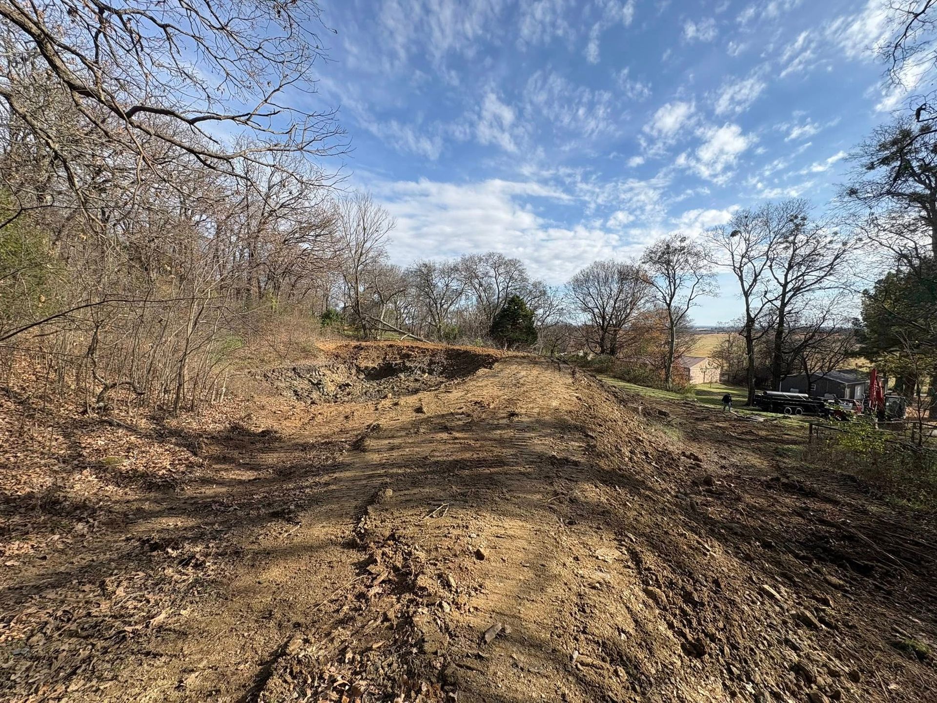 Dirt path through a wooded area on a sunny day. Trees with bare branches line the sides.