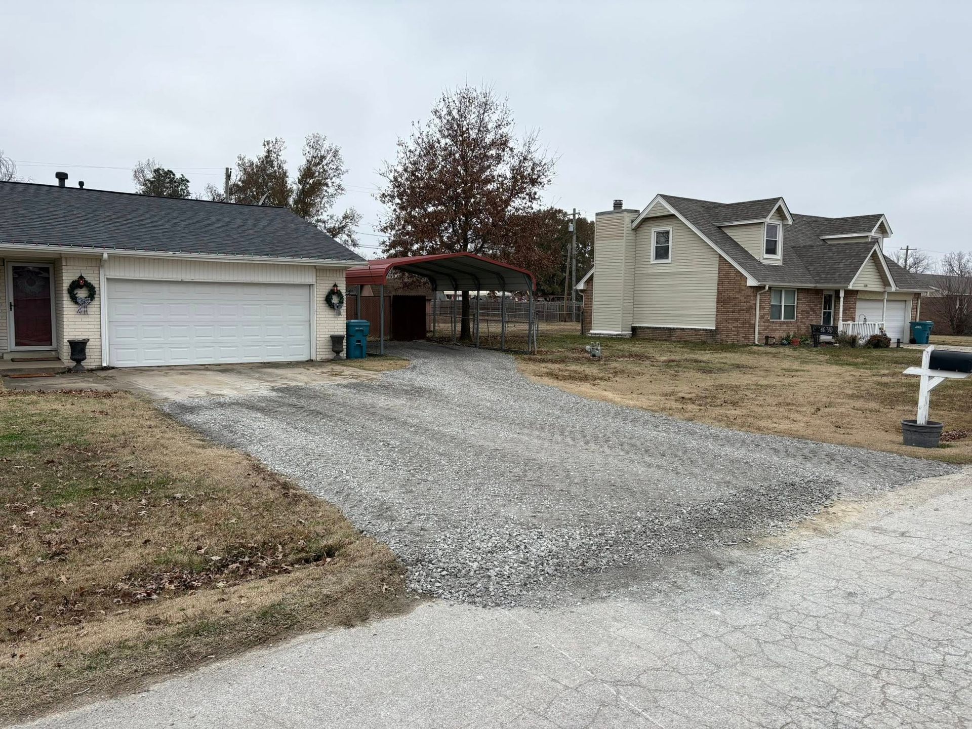 Gravel driveway in front of a house. Another house is visible to the right. Overcast sky.