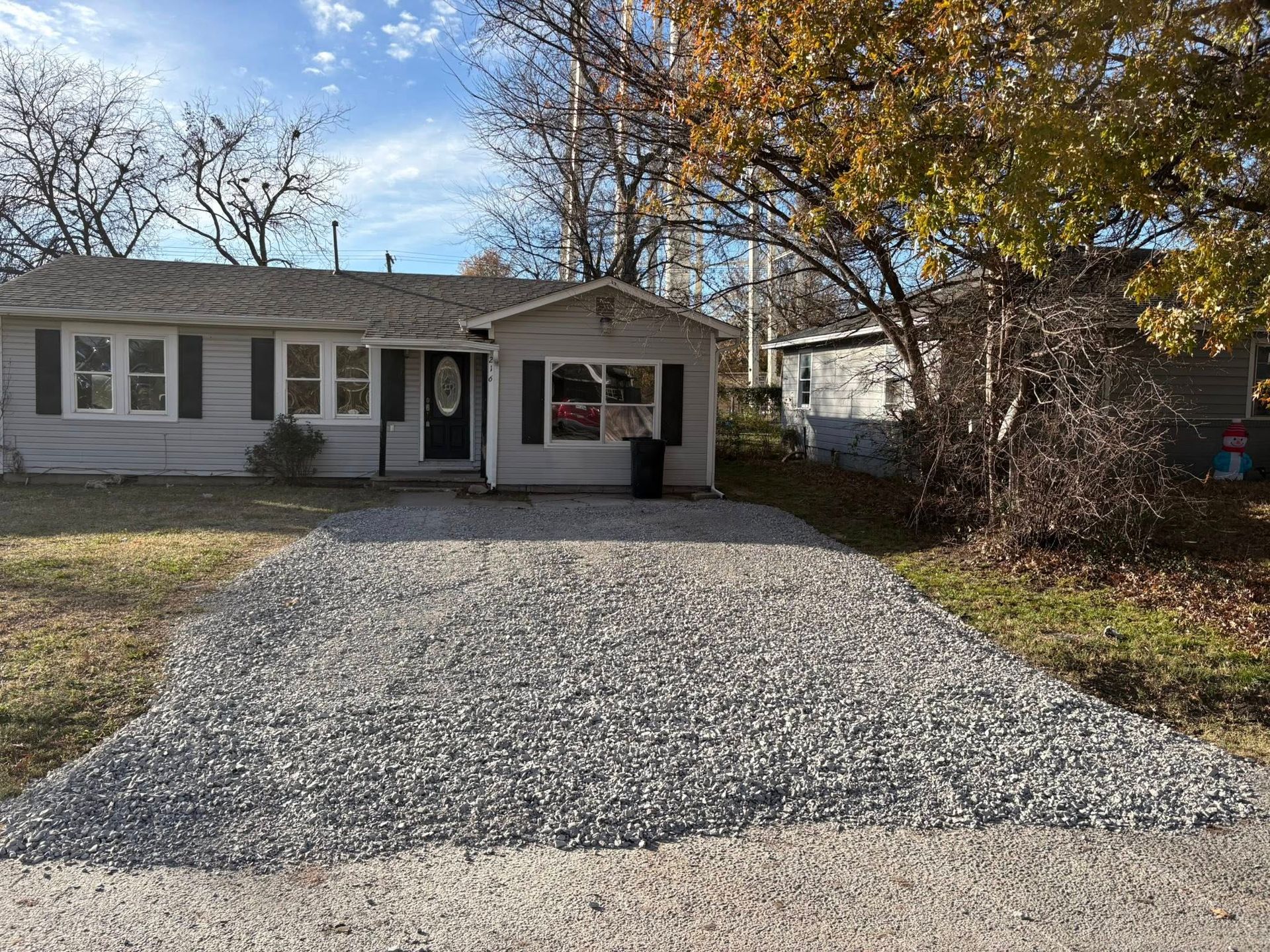 Gravel driveway leading to a small house with a black front door and gray siding. Autumn trees in the background.