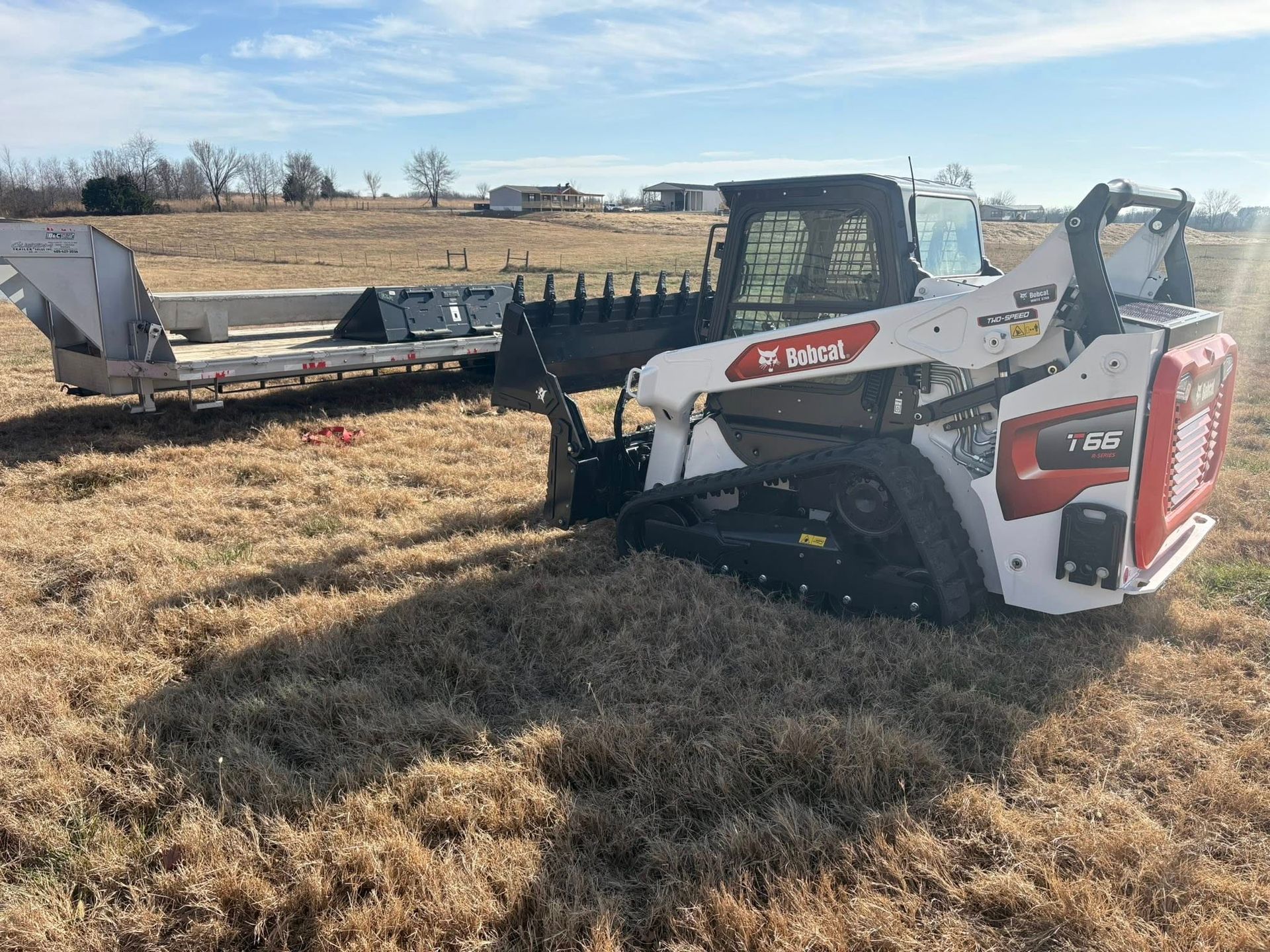 Bobcat T590 compact track loader with a rake attachment, on a grassy field, near a trailer.