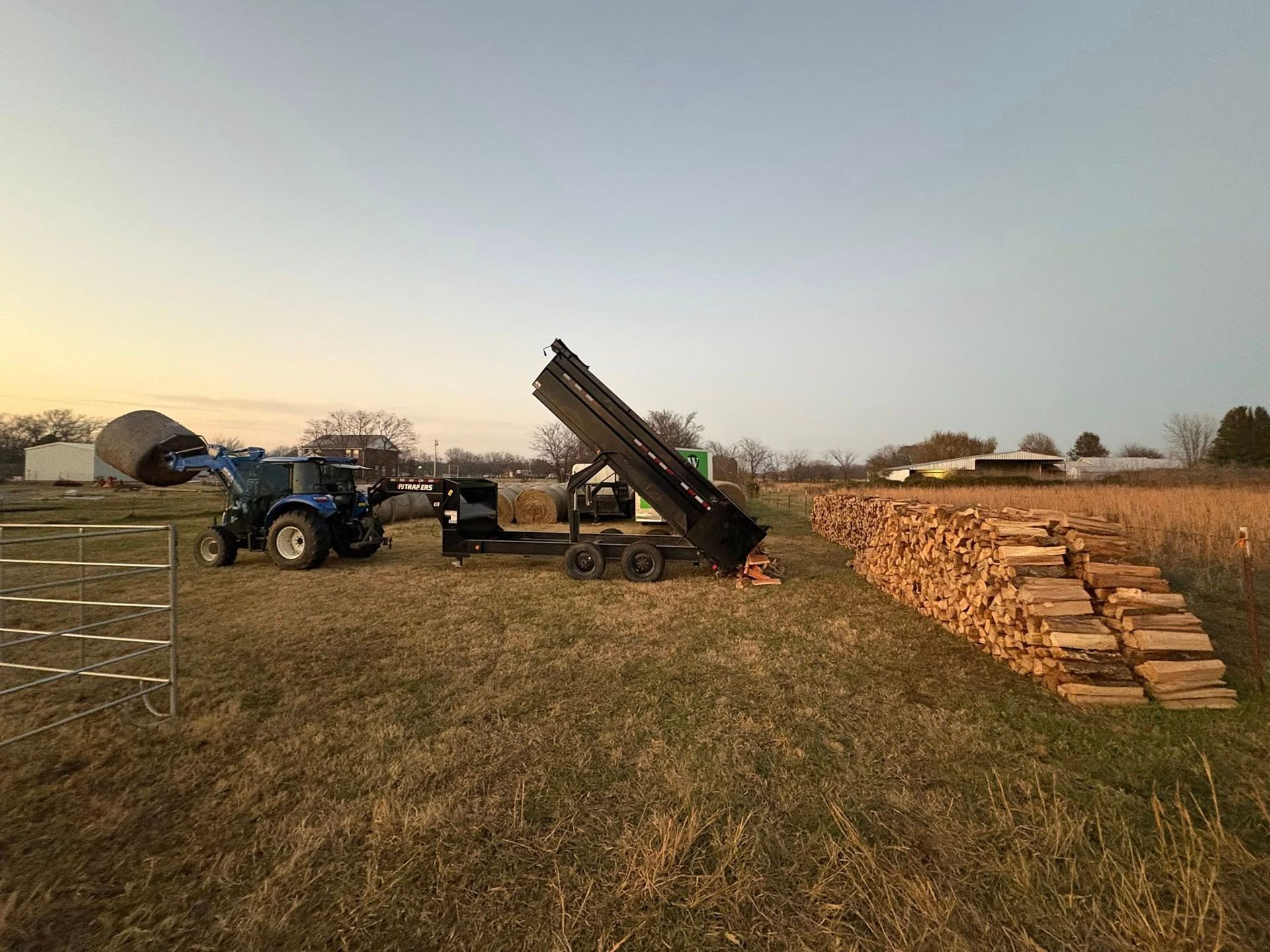 Blue tractor and dump trailer with a load of wood, in a field at sunset.