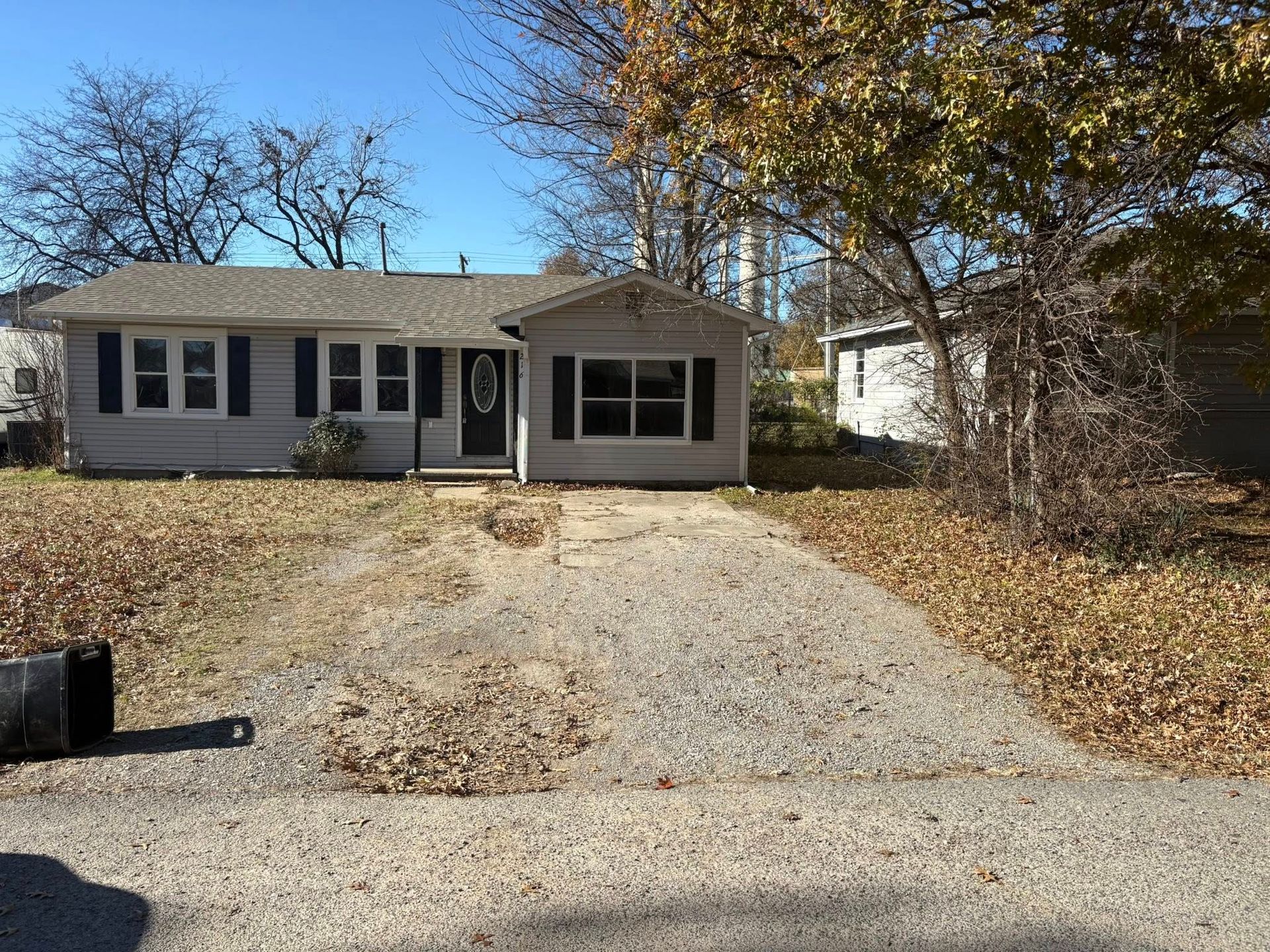 A small, weathered, one-story house with gravel driveway and fall foliage.
