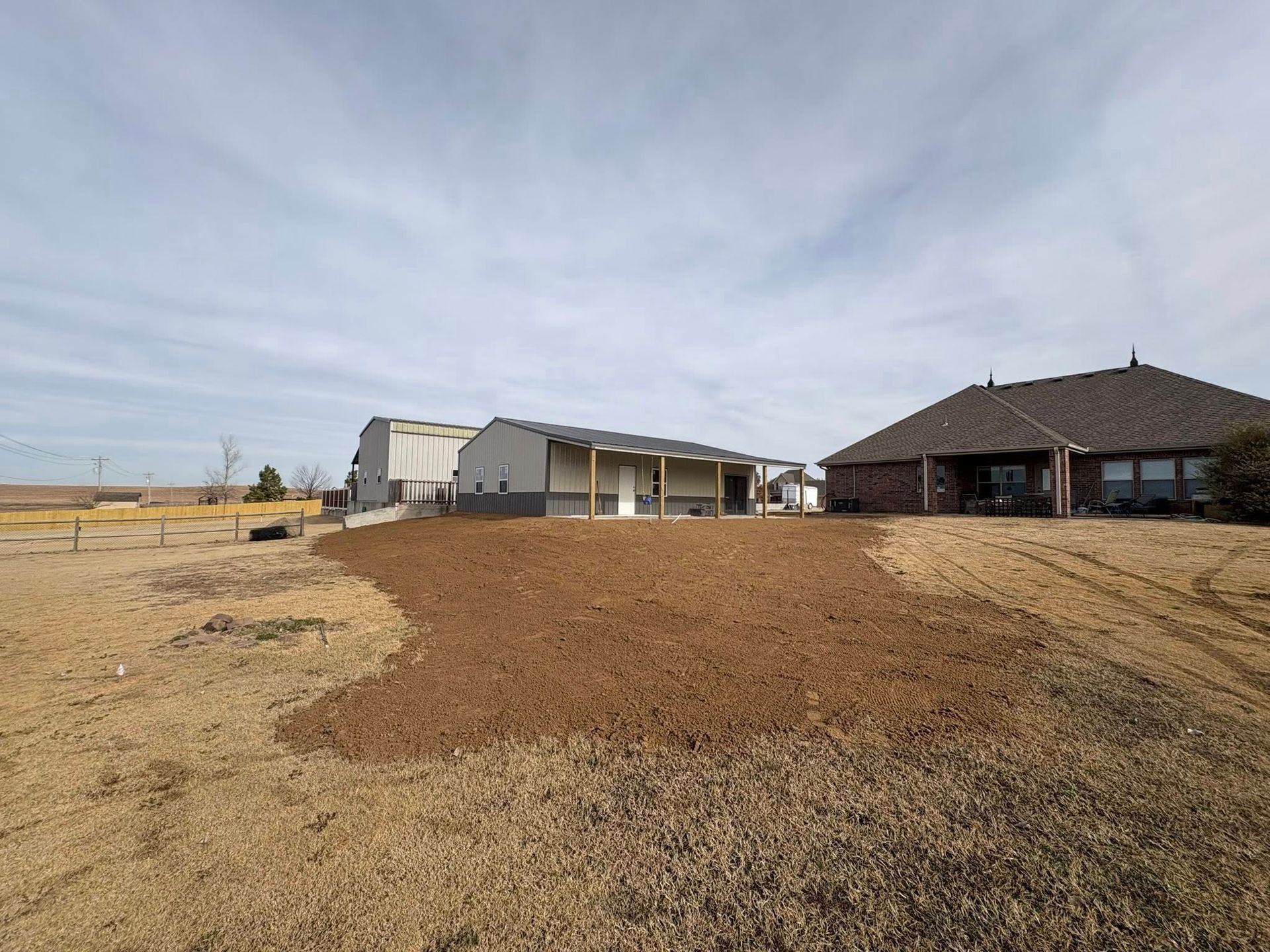 A backyard with a newly tilled area of soil in front of a house, shed, and blue sky.