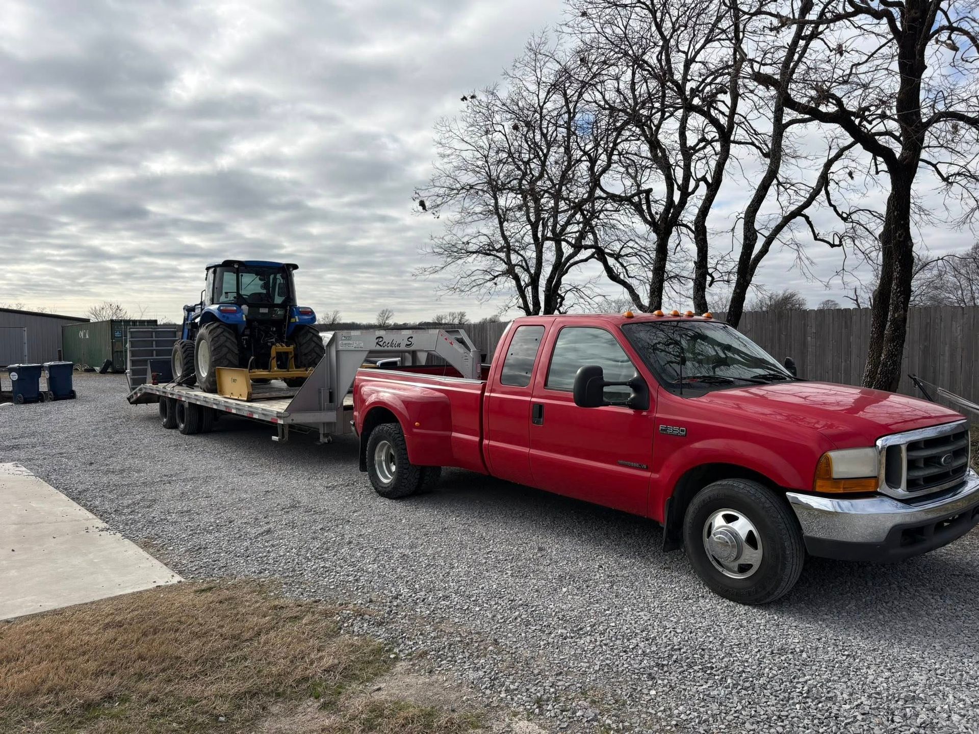 Red Ford truck pulling a trailer with a blue tractor on a gravel driveway under a cloudy sky.