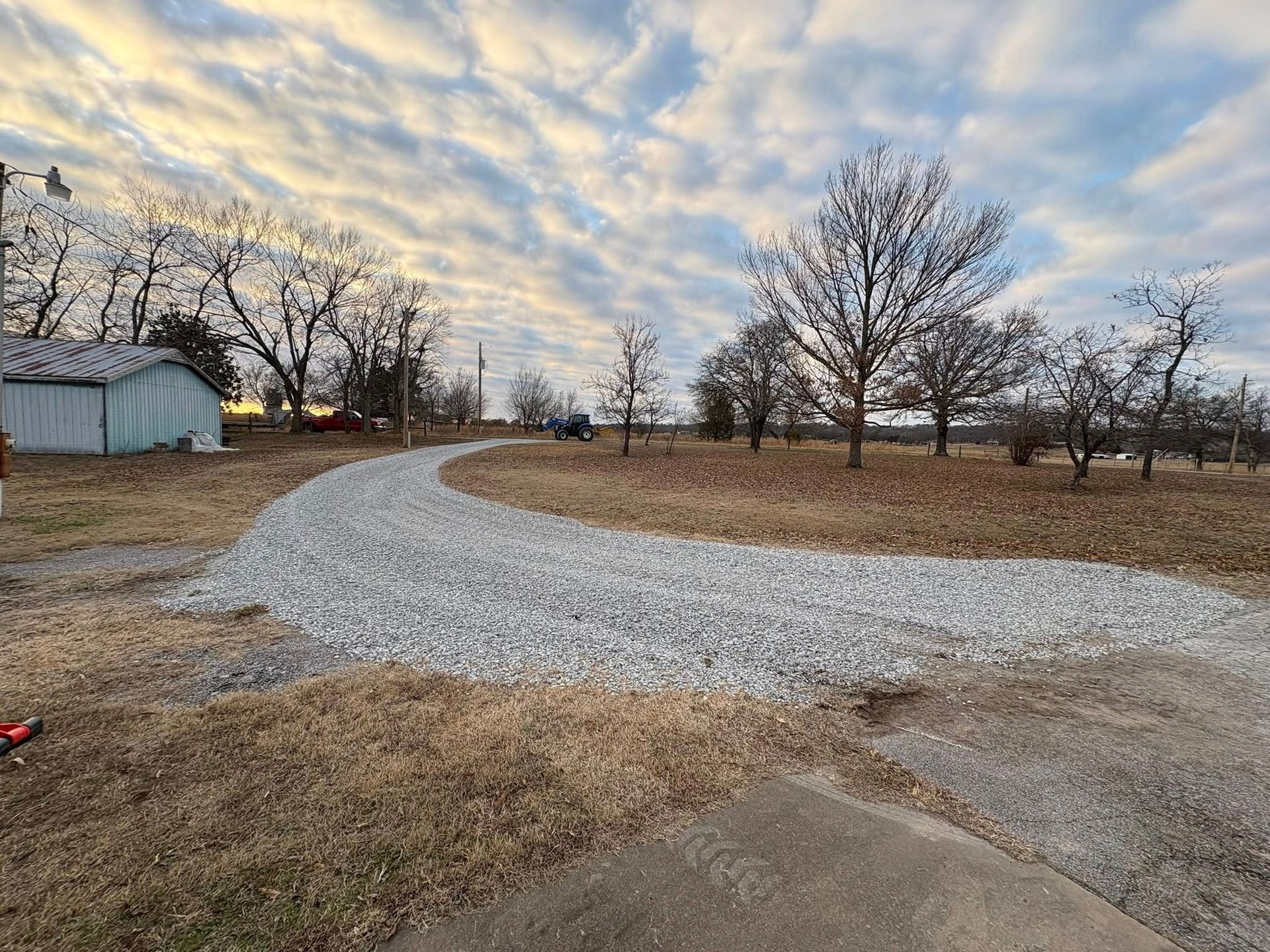 Gravel driveway curves through a brown field under a cloudy sky. Bare trees and a small blue building are visible.