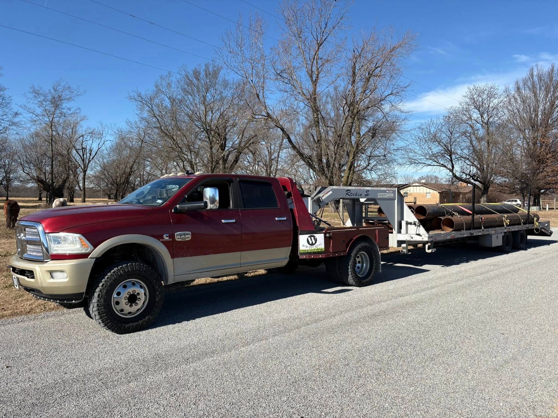 Red and tan pickup truck towing a trailer loaded with logs on a gravel road under a blue sky.