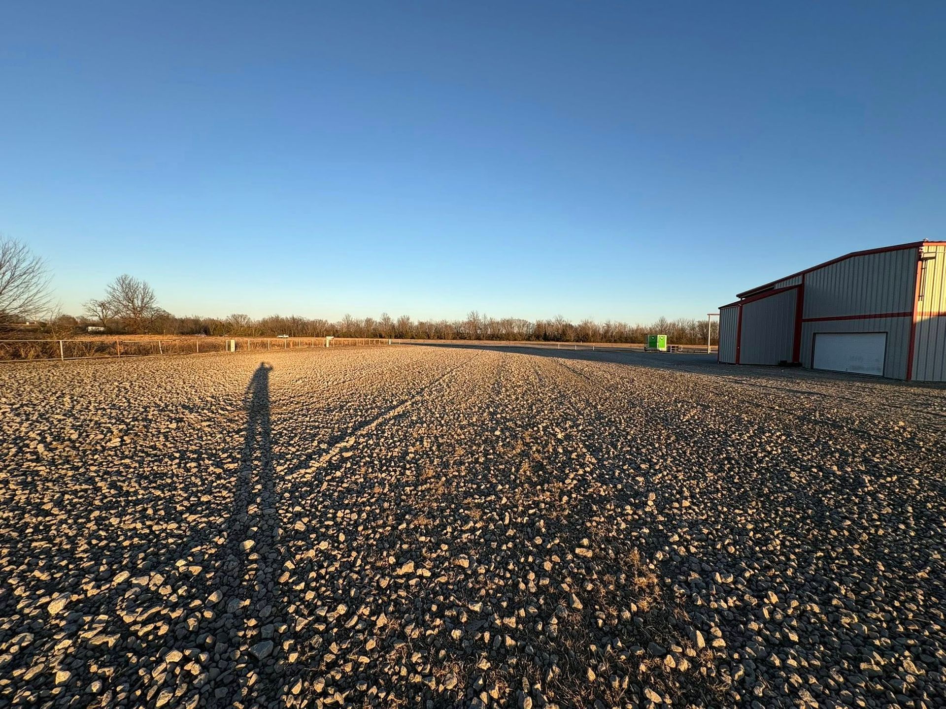 A gravel lot with a long shadow cast by the setting sun, a metal shed on the right, and a clear blue sky.