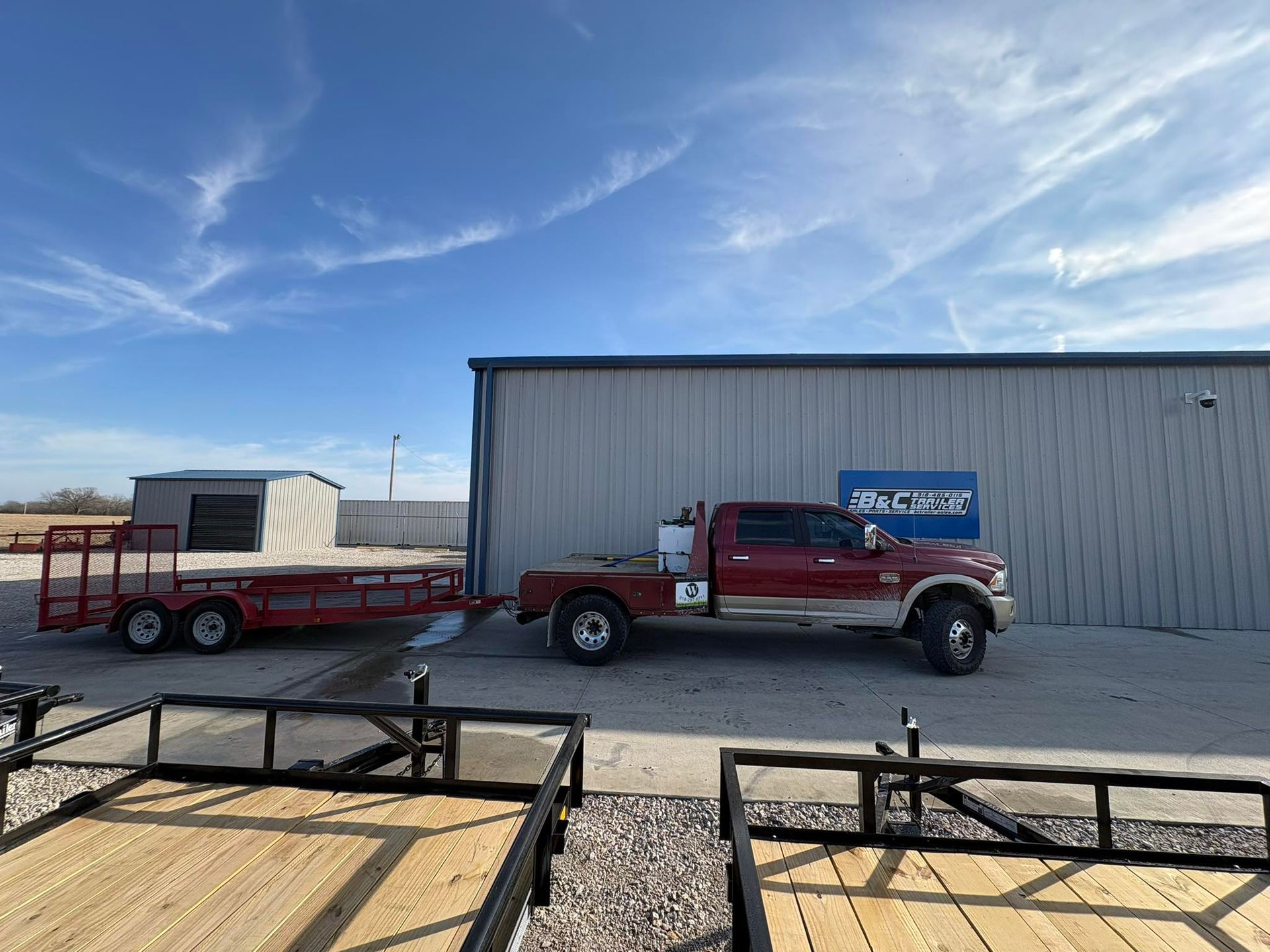 Red truck towing a red flatbed trailer, parked near a metal building on a sunny day.
