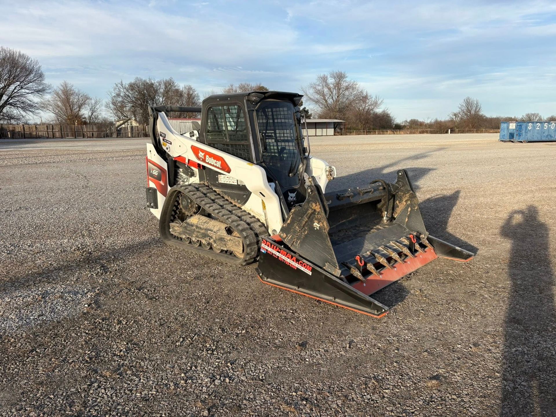 Bobcat skid-steer loader with a brush cutter attachment on a gravel lot under a bright sky.