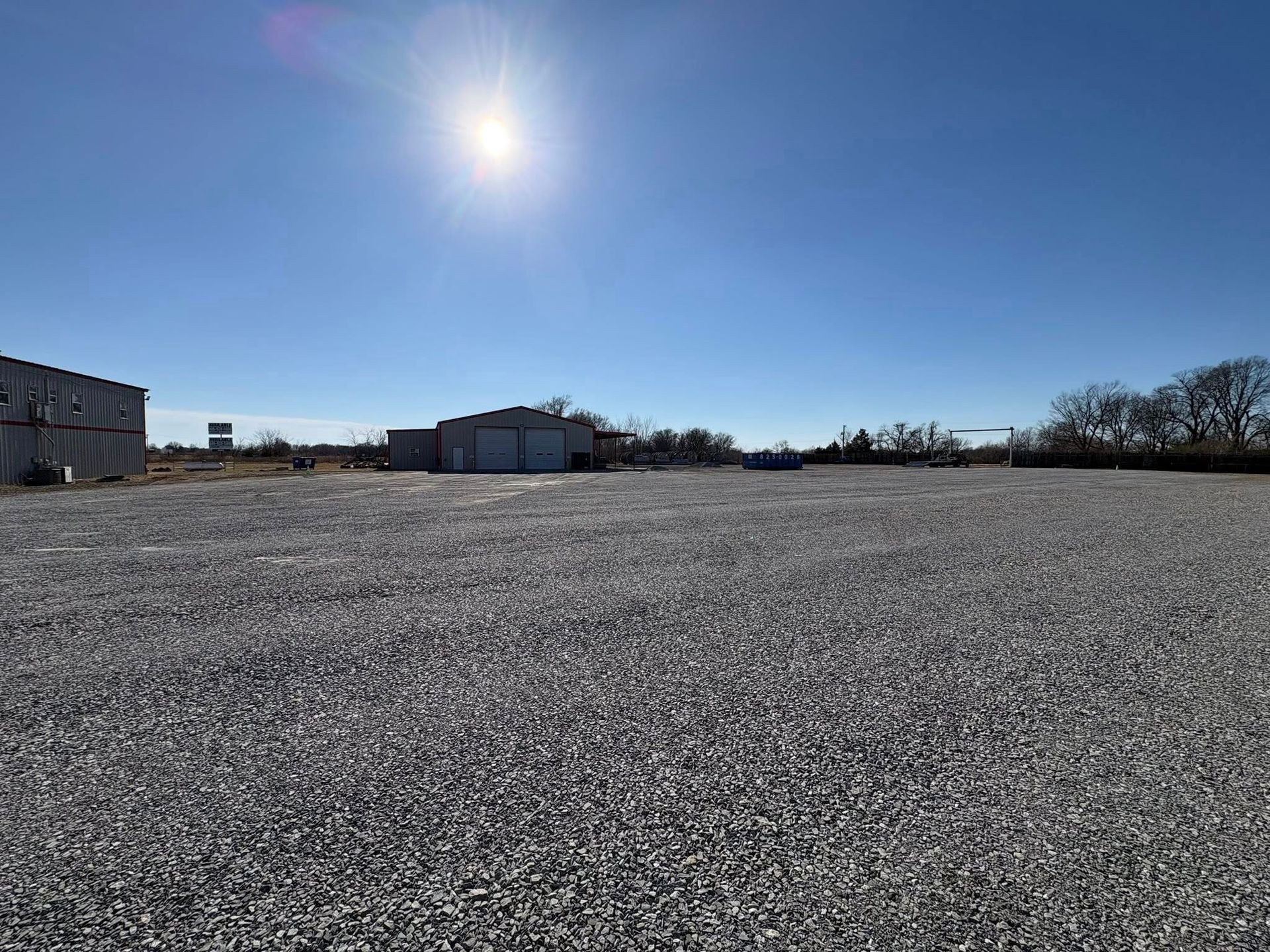 Gravel lot with buildings under a bright sun and blue sky.