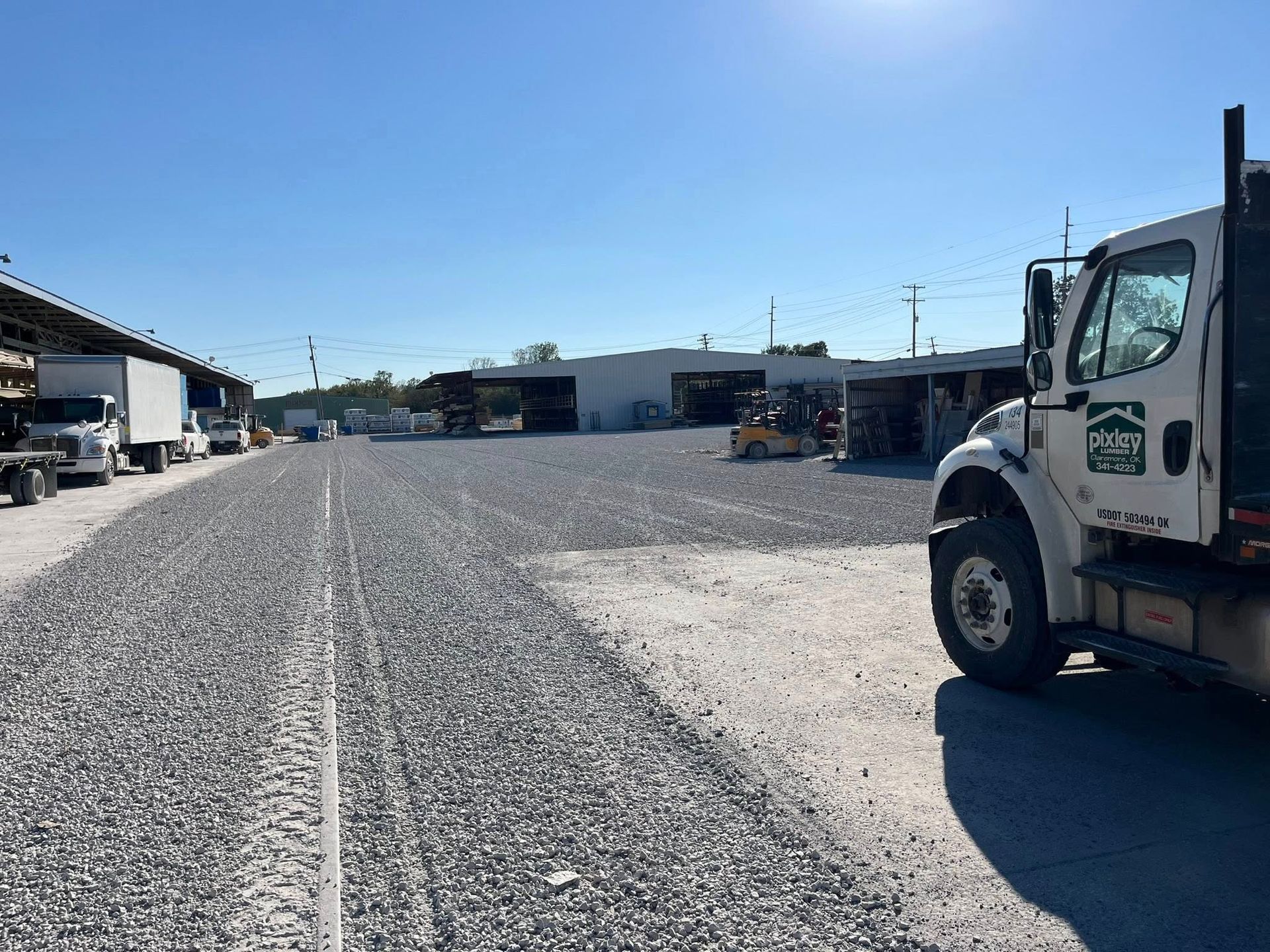 Gravel lot with trucks and warehouse buildings under a sunny sky.