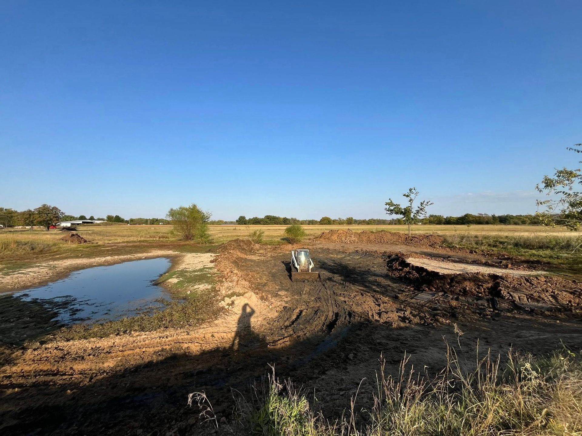 A tractor in a muddy field next to a pond under a clear blue sky.