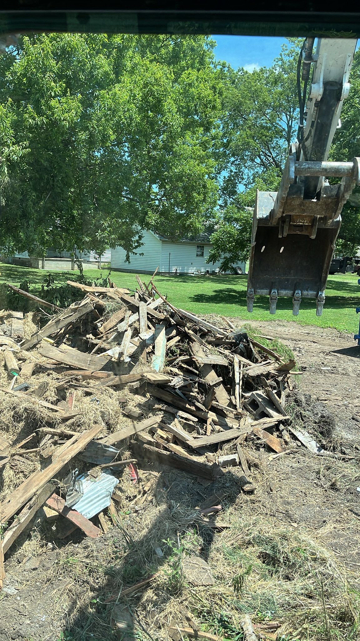 Excavator removing debris from a demolition site; wood and dirt piled up; green trees and a house in the background.