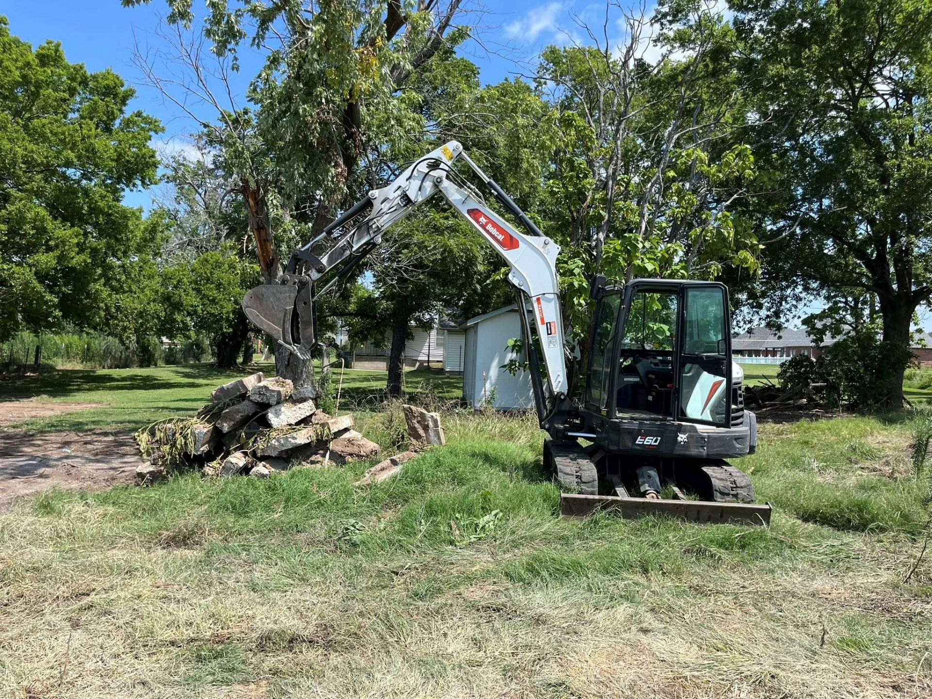 Small excavator with raised bucket, moving stone and debris outdoors near trees.